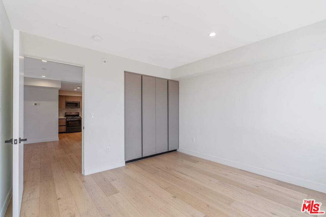 Vacant bedroom with light wood floors and a built-in sliding-door closet; kitchen visible through the doorway.