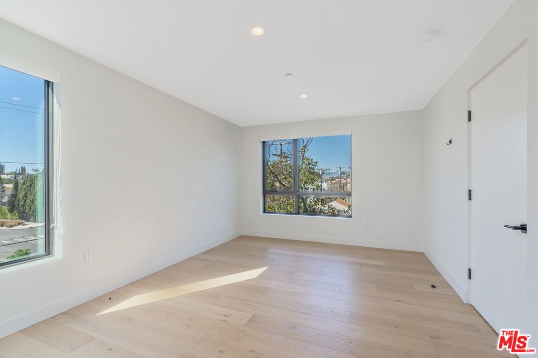 Vacant room with pale wood floors, white walls, and windows overlooking nearby buildings and trees.