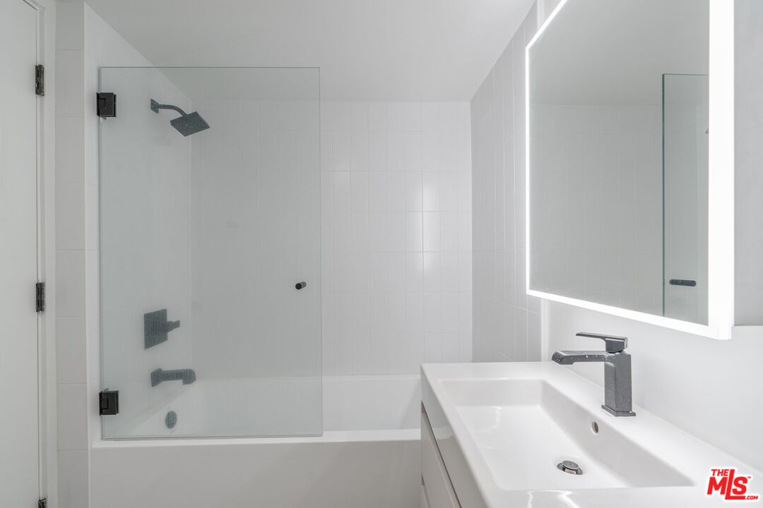 Modern bathroom with a glass-enclosed tub, dark shower fixtures, and an illuminated mirror above the vanity.
