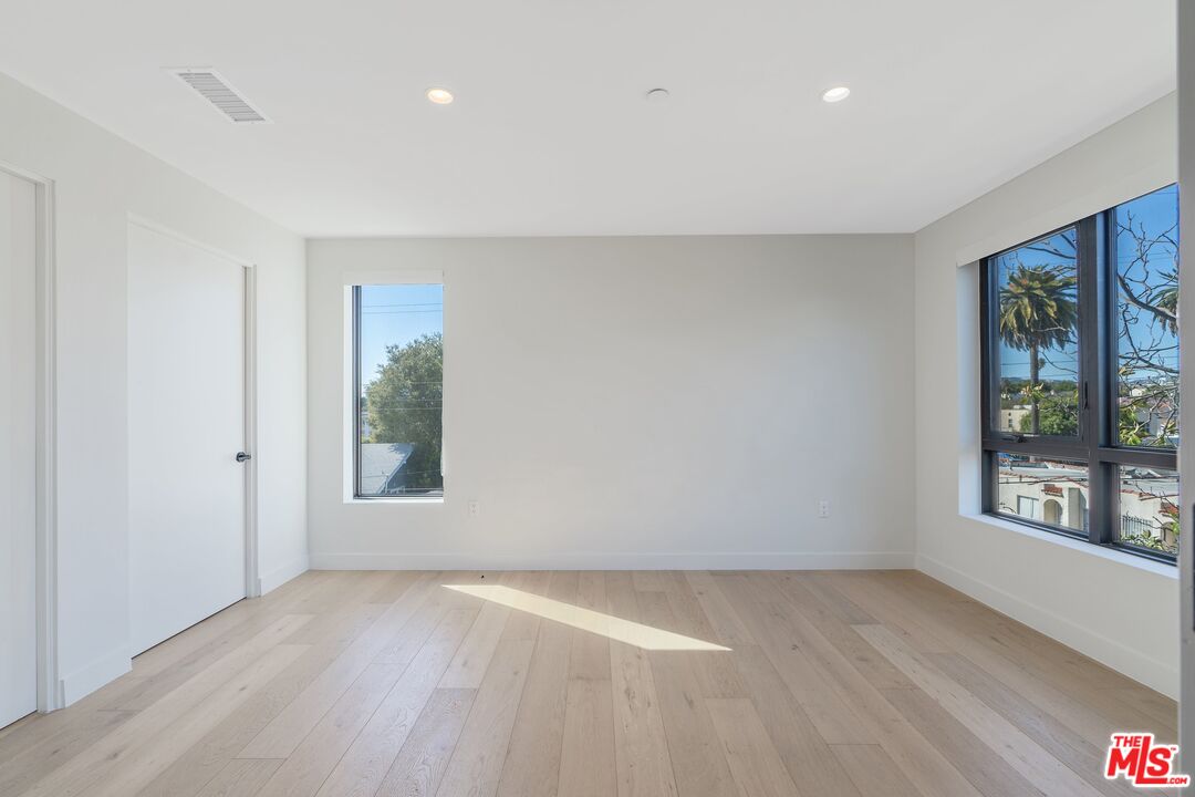 Bright vacant room with pale wood floors and large windows overlooking palm trees and nearby homes.