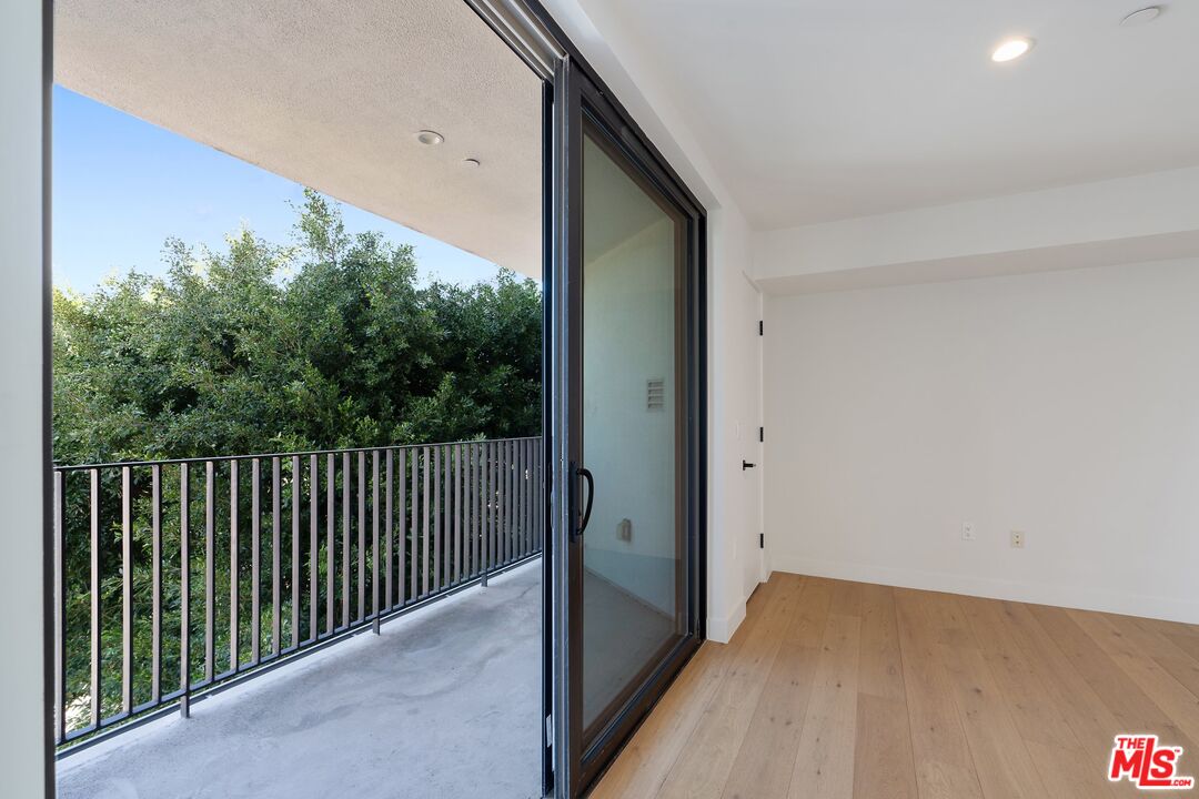 Empty room with light wood floors and a sliding glass door to a leafy balcony.