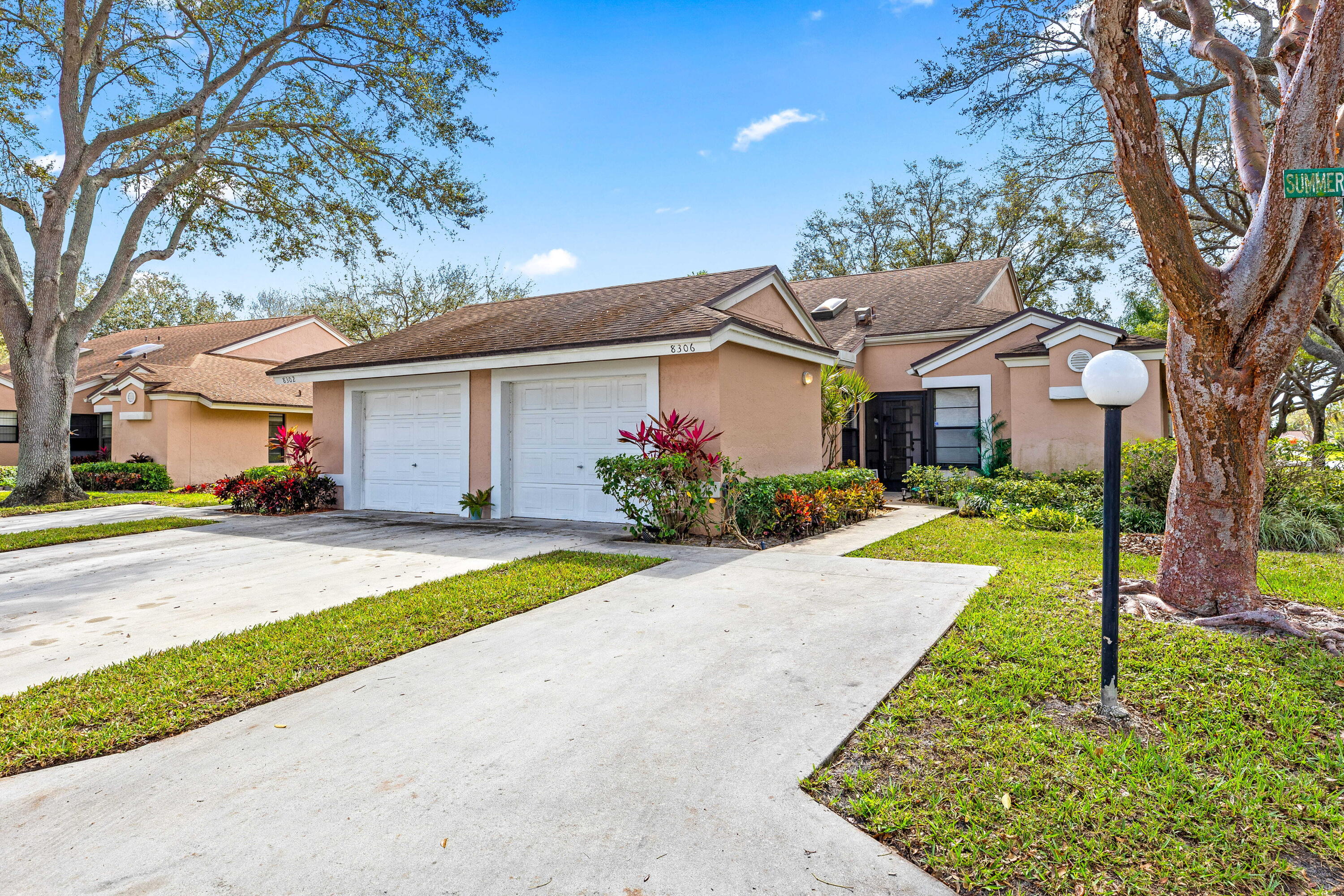 Peach single-story home with two white garage doors, landscaped shrubs, and a broad driveway.