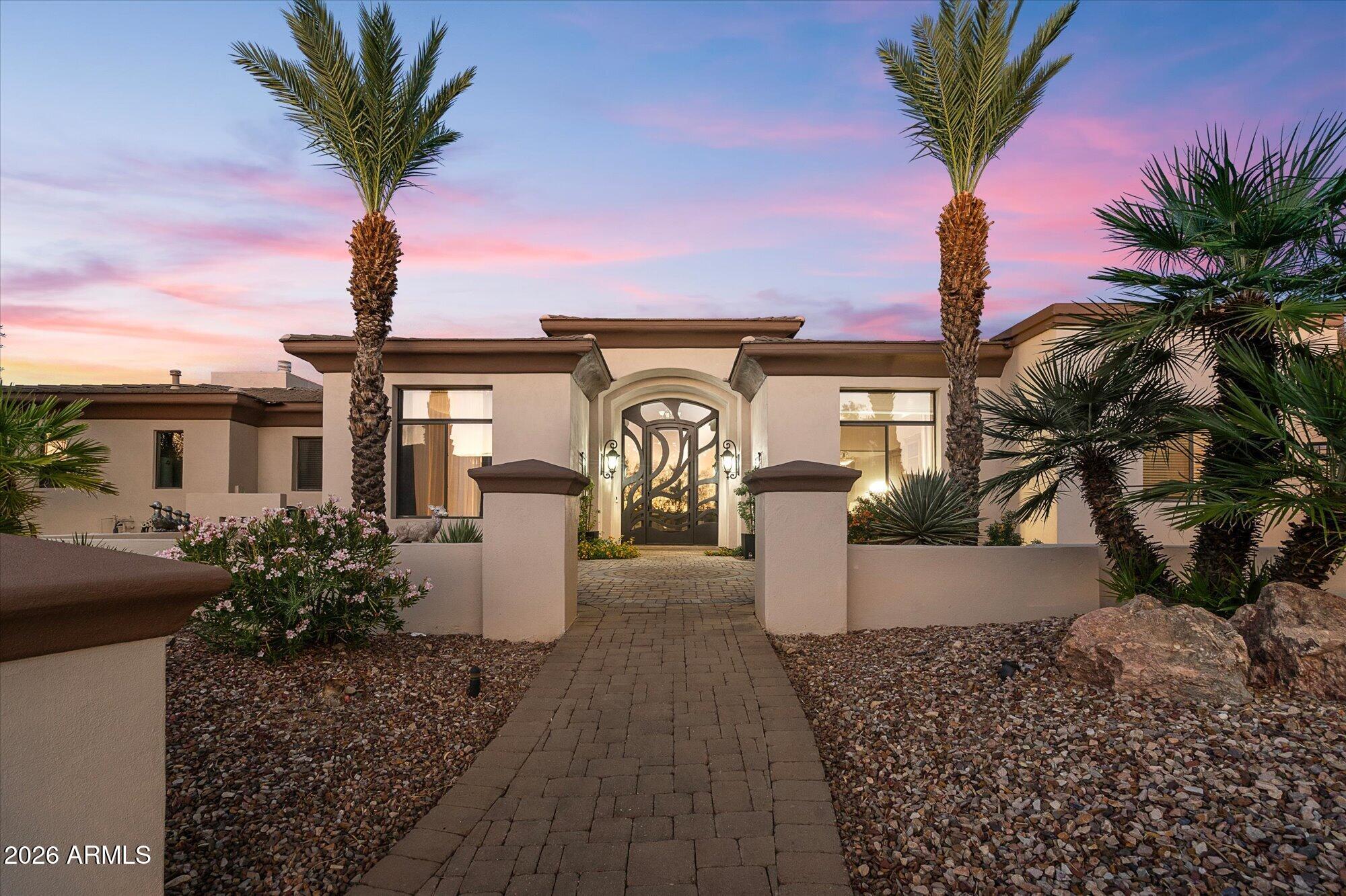Single-story home with an ornate entry gate, paver walkway, desert landscaping, and palms at dusk.