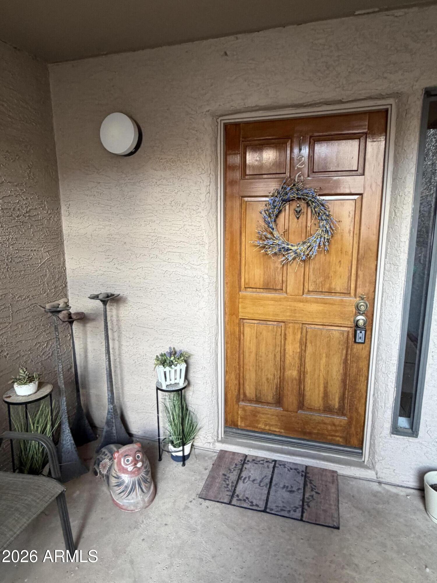 Stucco home entry with a paneled wooden door, wreath, potted plants, and welcome mat.
