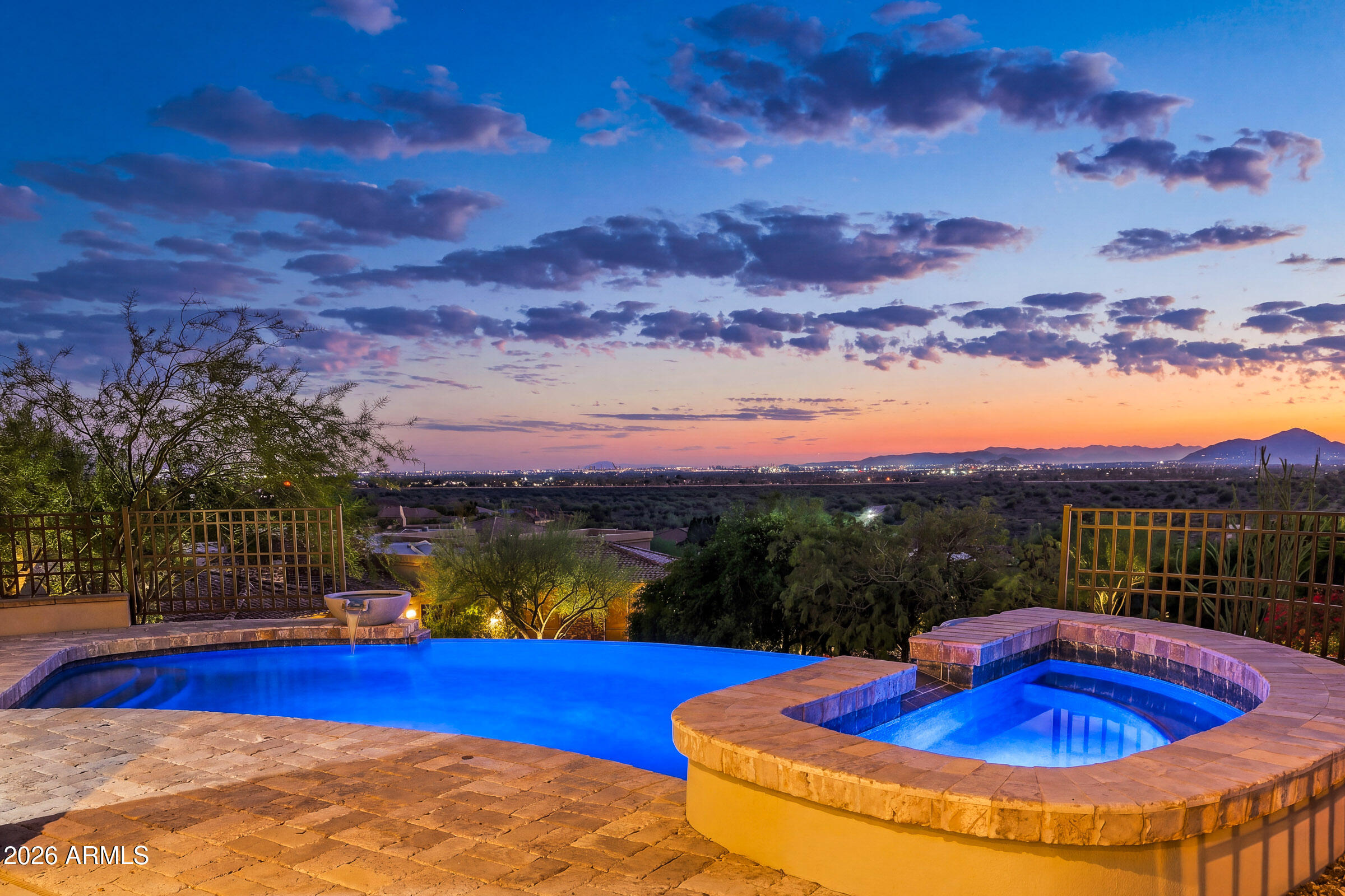 Pool and raised spa overlook desert vegetation, city lights, and distant mountains at dusk.