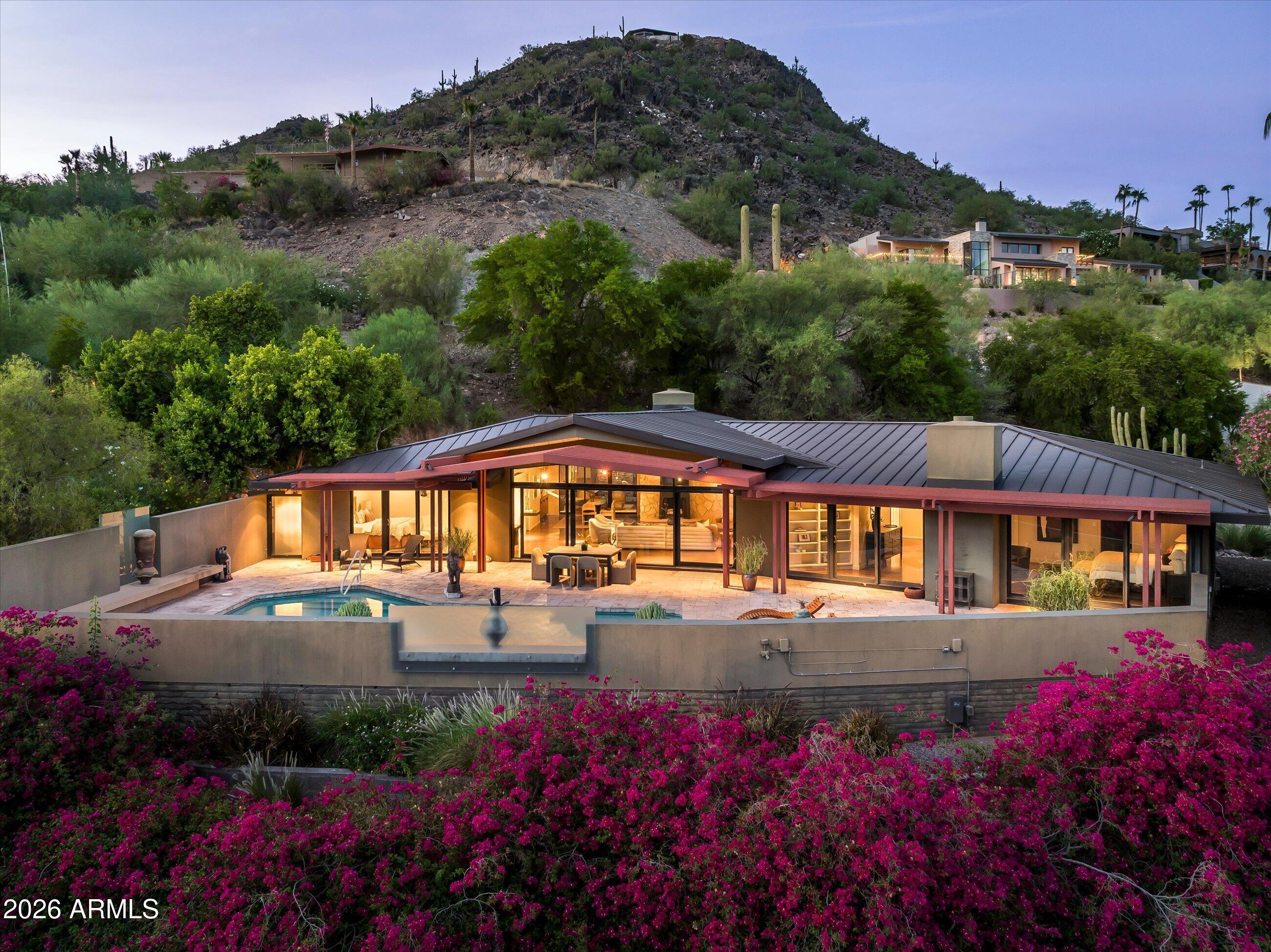Modern home with glass walls and pool below a cactus-covered hillside, surrounded by bright magenta flowers at dusk.