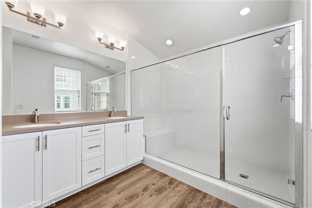 Bathroom with a double-sink vanity, white cabinetry, glass-enclosed shower, and wood-look flooring.