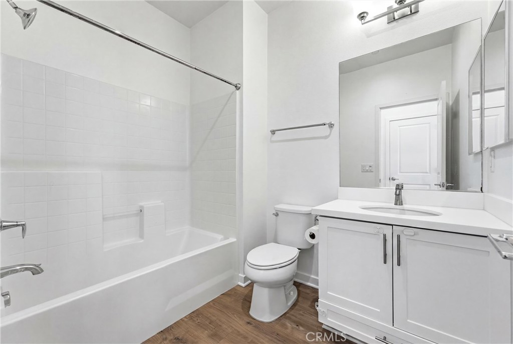 Bathroom with a white tiled tub and shower, toilet, and vanity over wood-look flooring.
