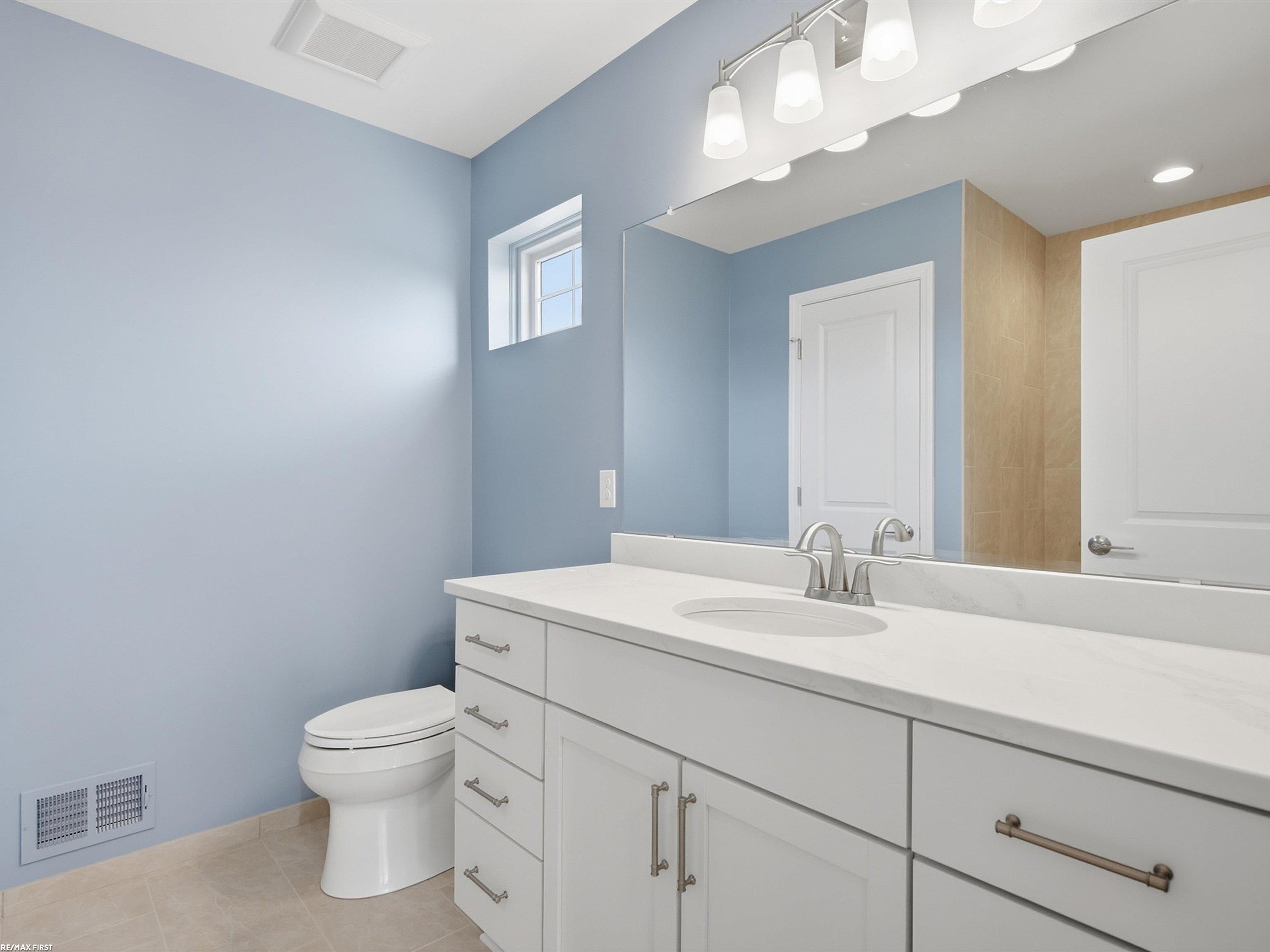 Bathroom with pale blue walls, a white vanity, large mirror, and small high window.
