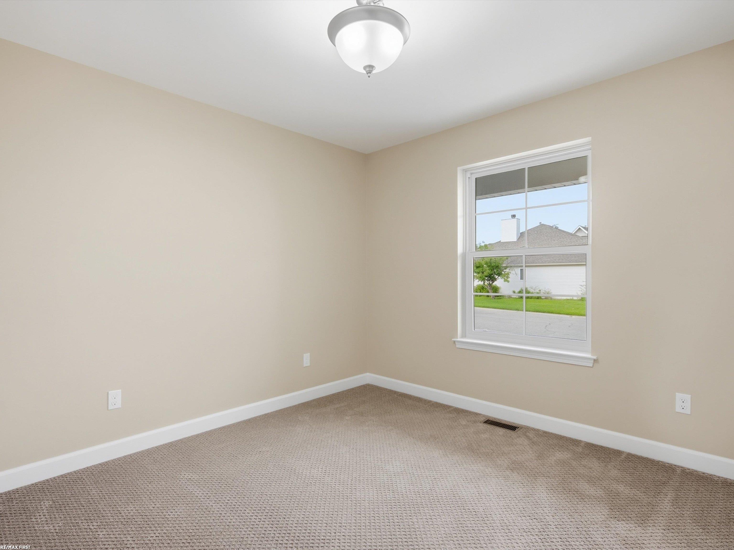 Vacant bedroom with beige walls, carpeted floor, and a window overlooking a neighboring house.
