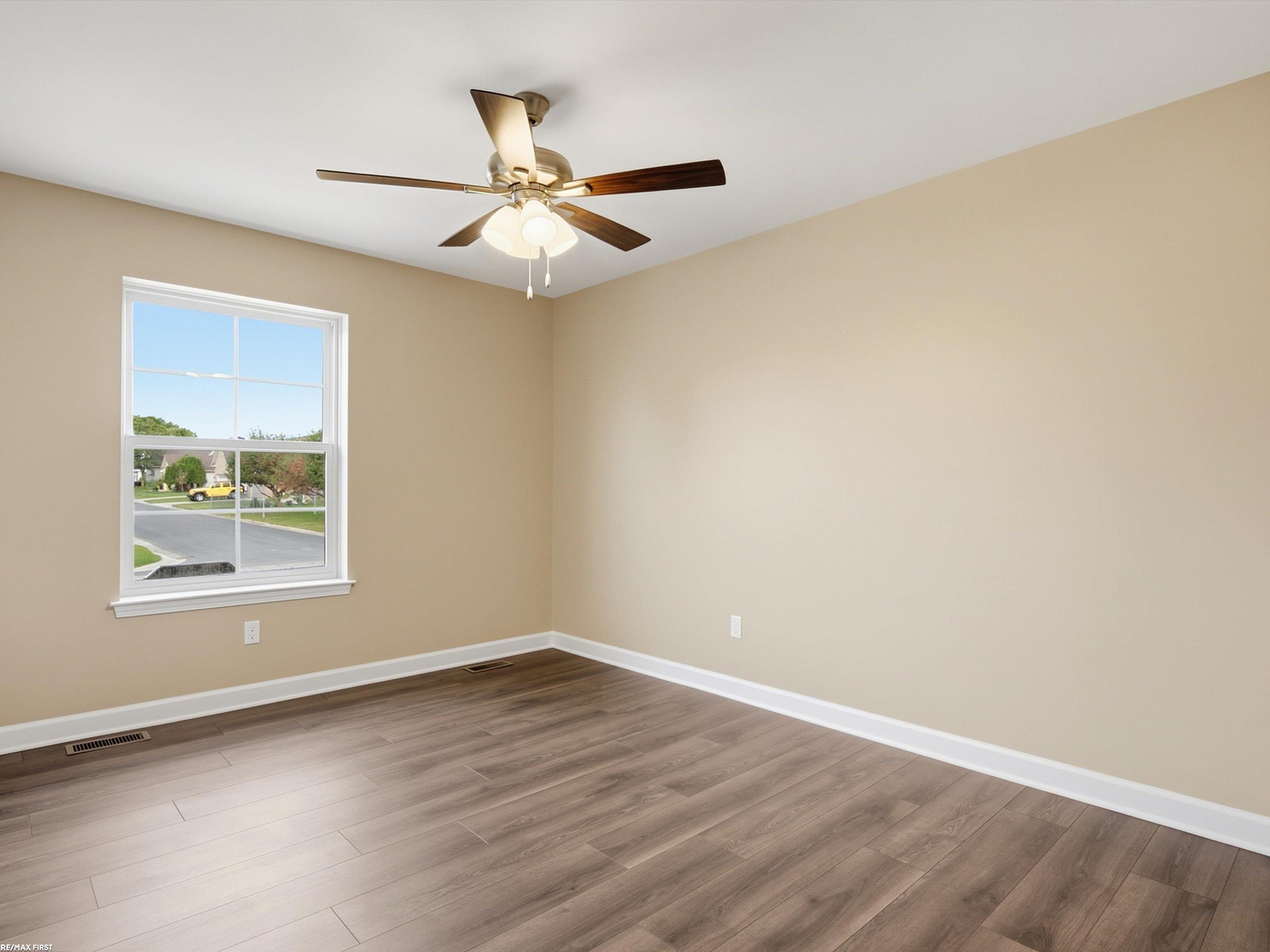 Empty room with plank flooring, beige walls, and a ceiling fan beside a window overlooking the street.