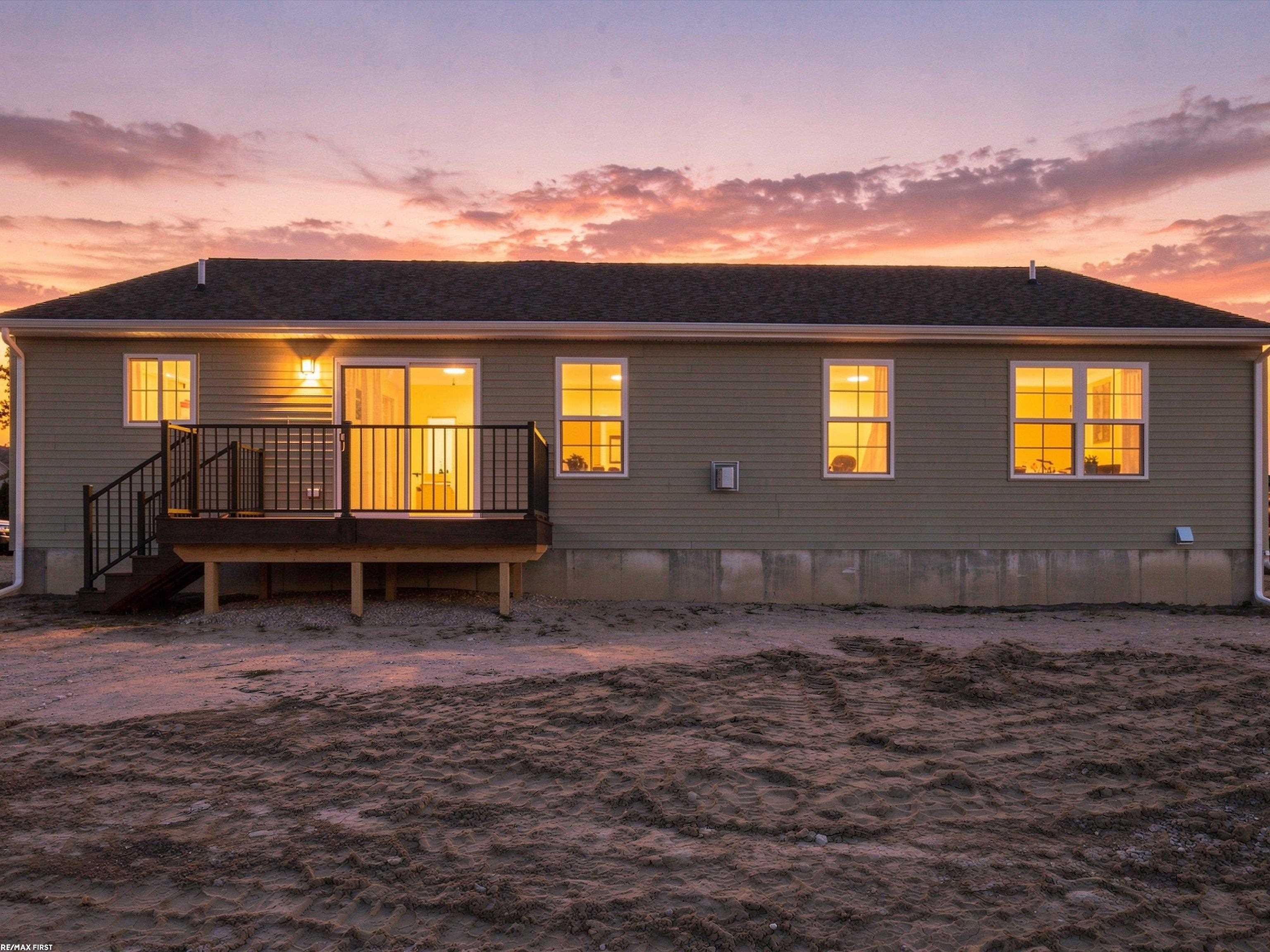 Rear of a gray-sided house at sunset, with a raised deck and warmly lit windows.