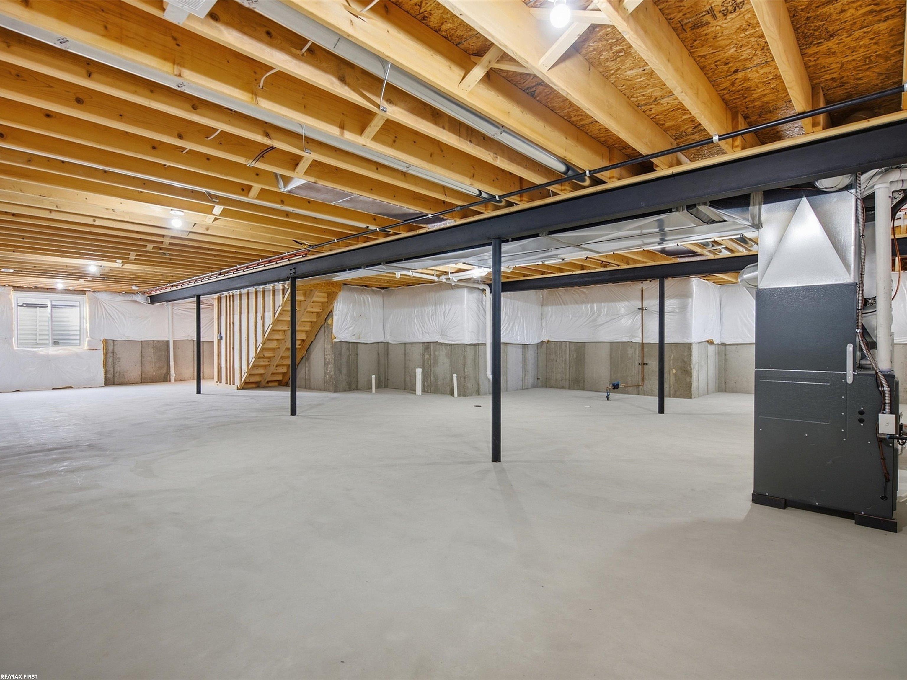 Unfinished basement with exposed ceiling joists, black support beams, concrete floor, stairs, and HVAC equipment.
