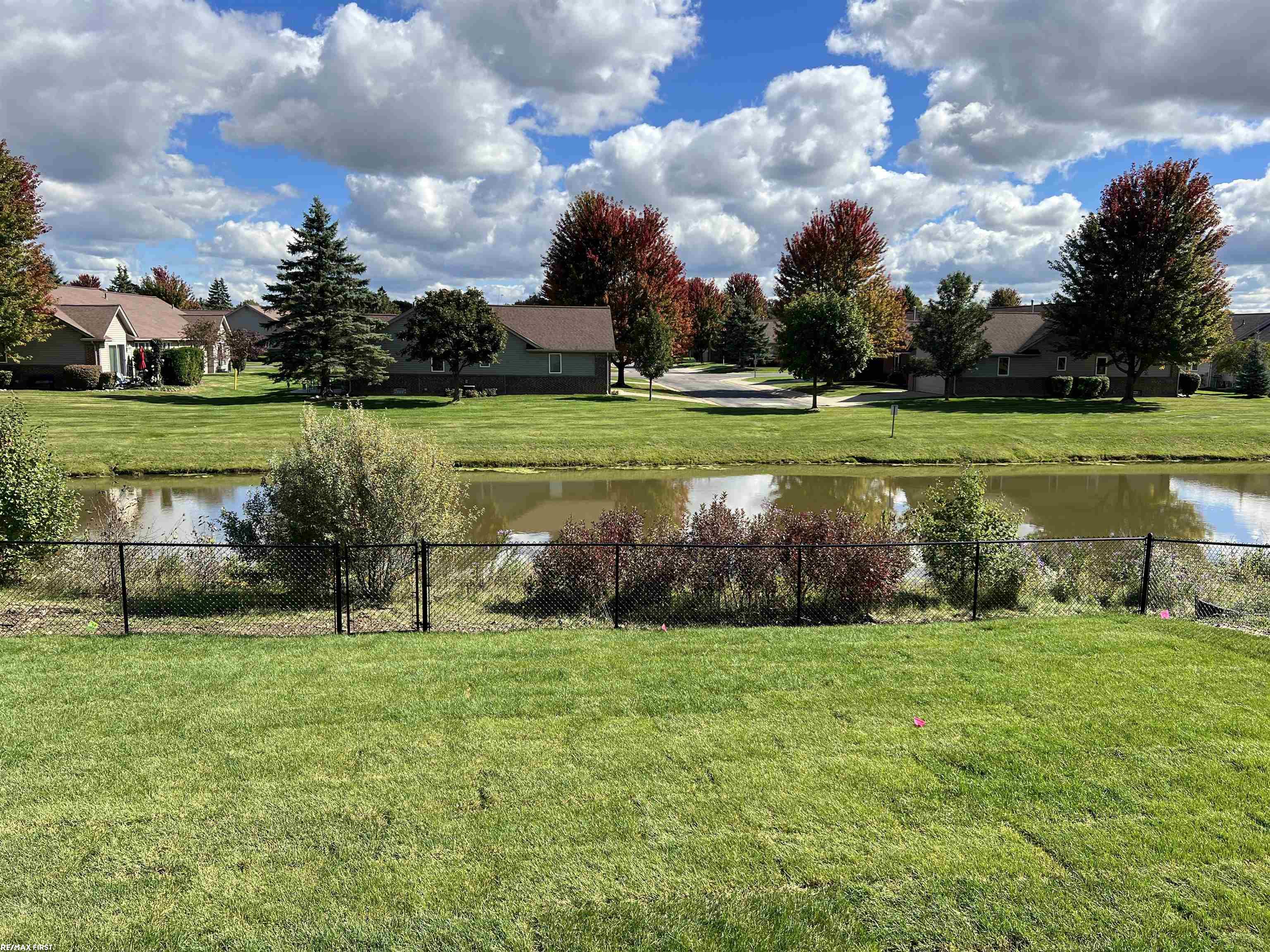 Pond beyond a fenced grassy lawn, with homes and autumn trees along the far bank.