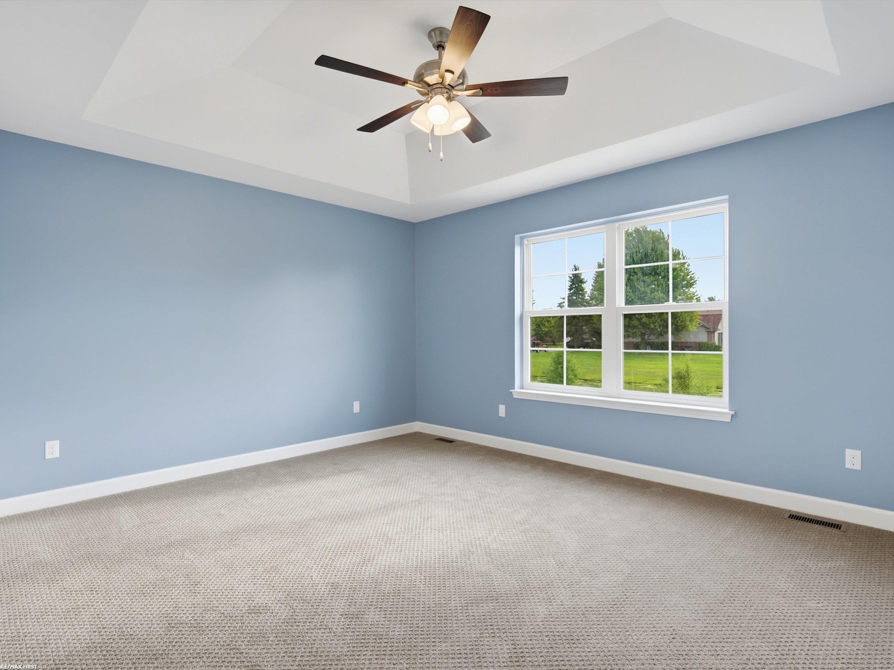 Empty blue bedroom with beige carpet, tray ceiling, ceiling fan, and a large window overlooking a lawn.