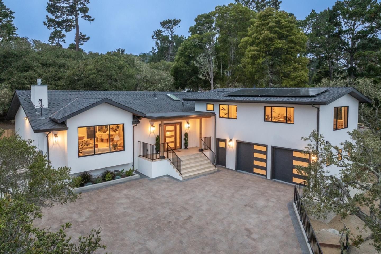 Modern white home with solar panels, a broad driveway, and mature trees at dusk.