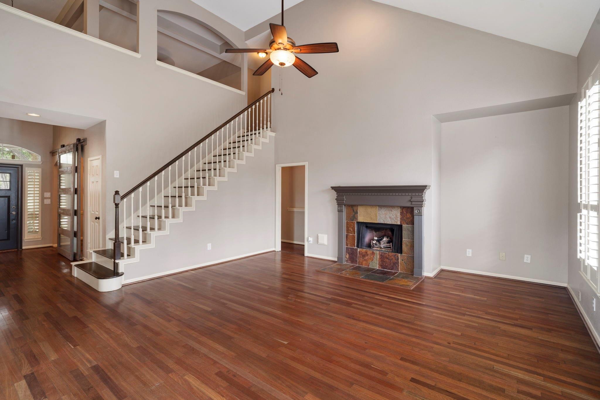 Unfurnished living room with hardwood floors, a stone fireplace, and a staircase beneath a vaulted ceiling.