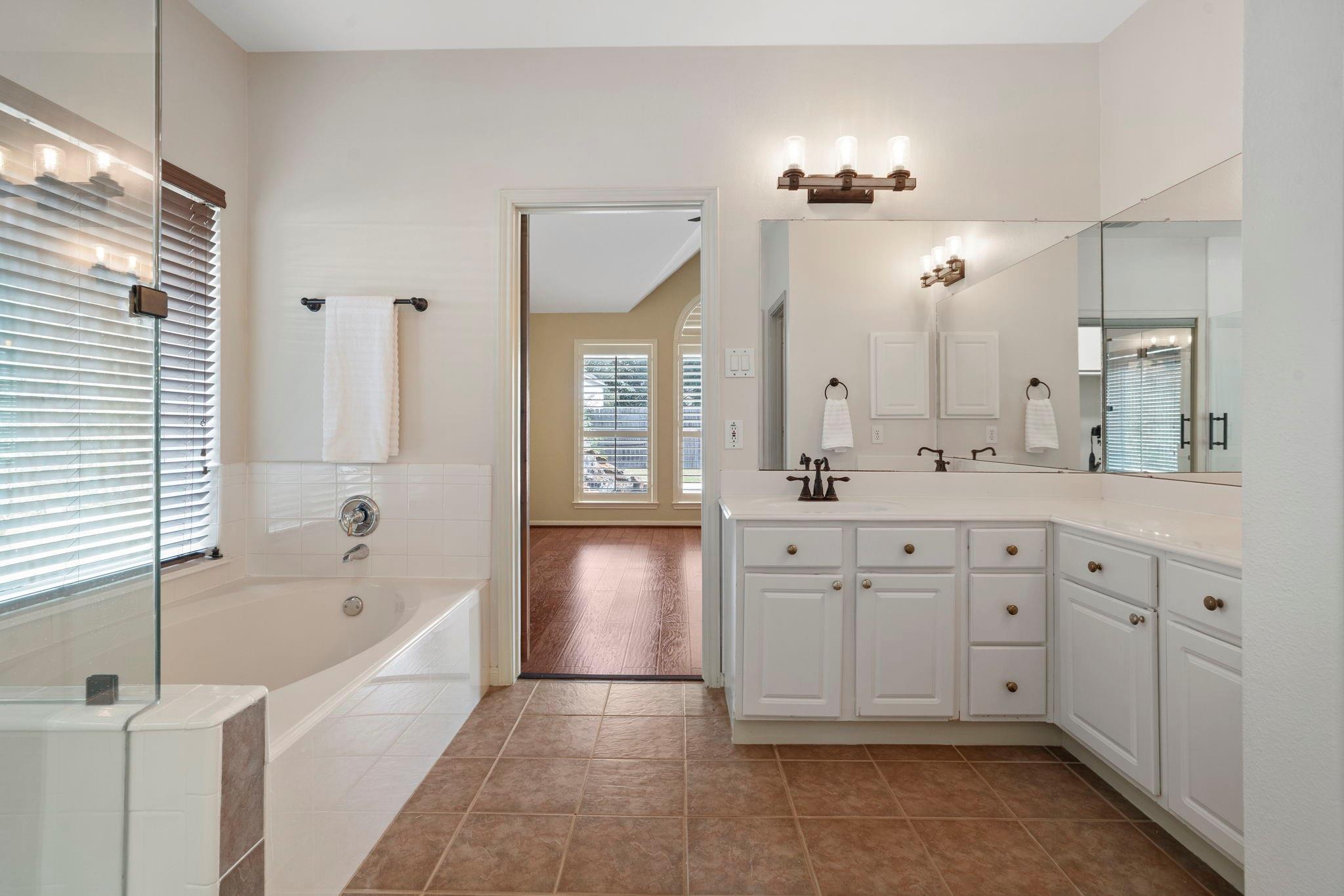 Bathroom with a glass-enclosed tub, long white vanity, large mirrors, and brown tile flooring.