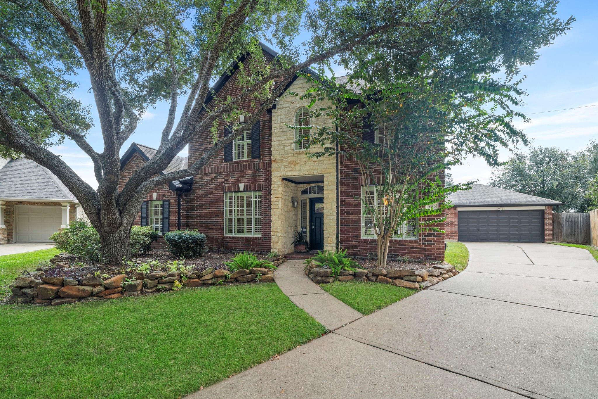 Two-story brick home with a stone entrance, mature oak, landscaped front lawn, and attached garage.