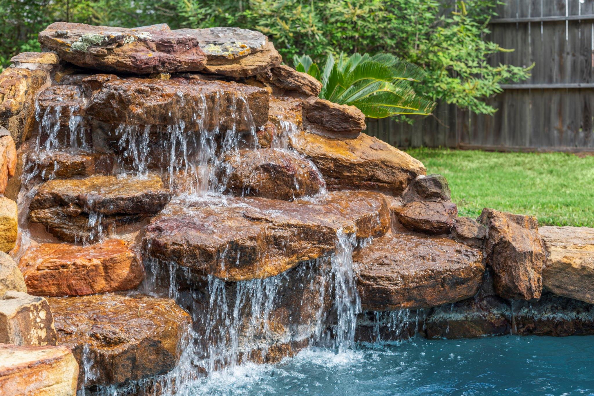 Backyard pool with water cascading over a broad, multilevel natural-rock waterfall.