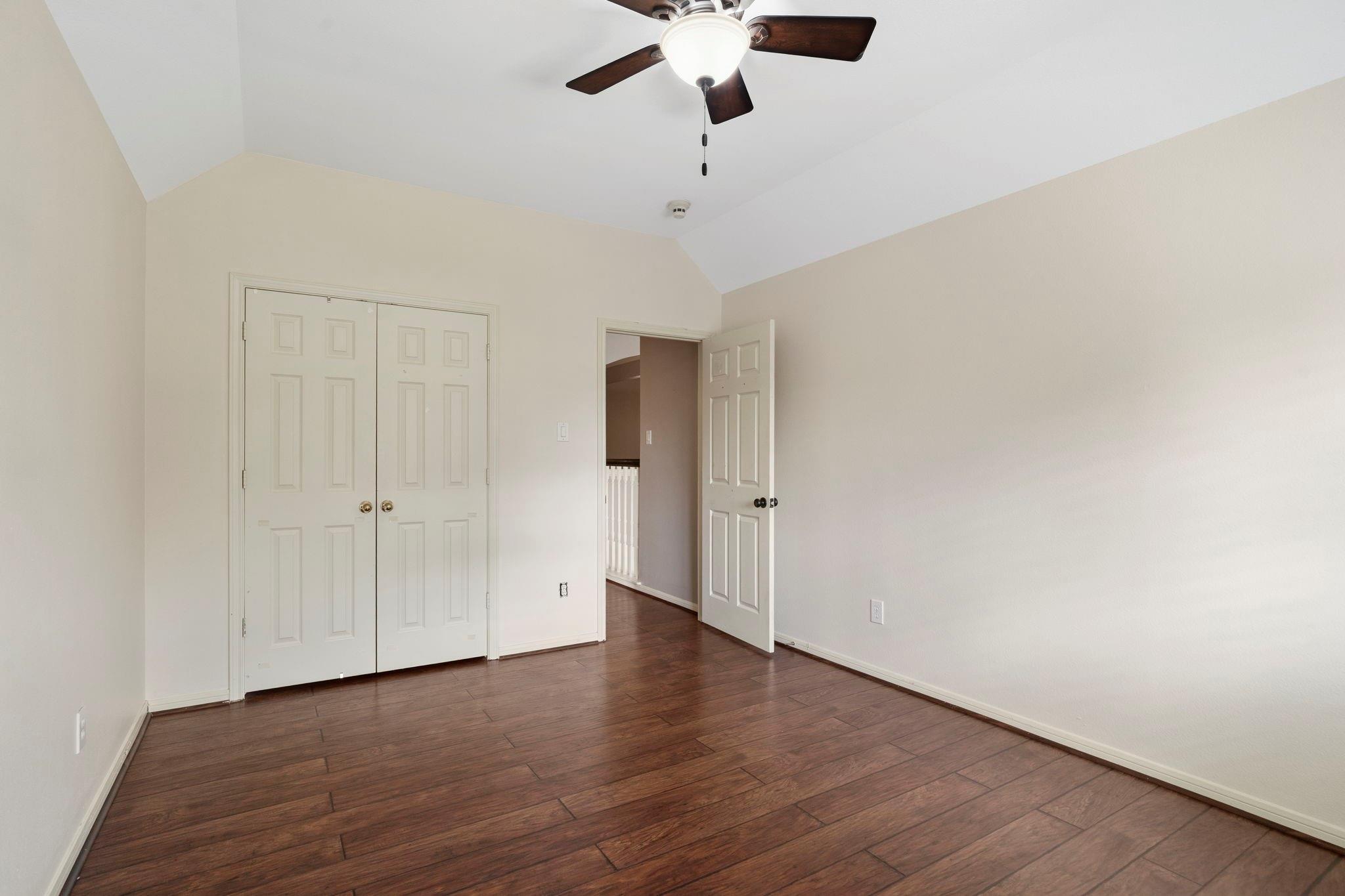 Empty bedroom with vaulted ceiling, dark plank flooring, double closet doors, and ceiling fan.