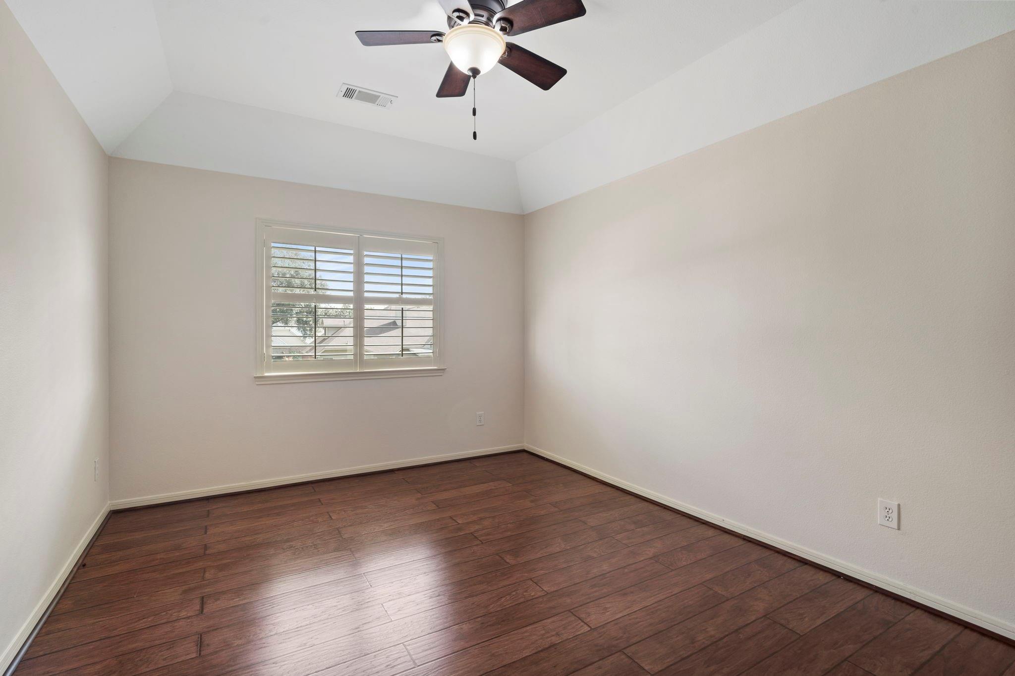 Empty room with dark plank flooring, vaulted ceiling, ceiling fan, and shuttered window.