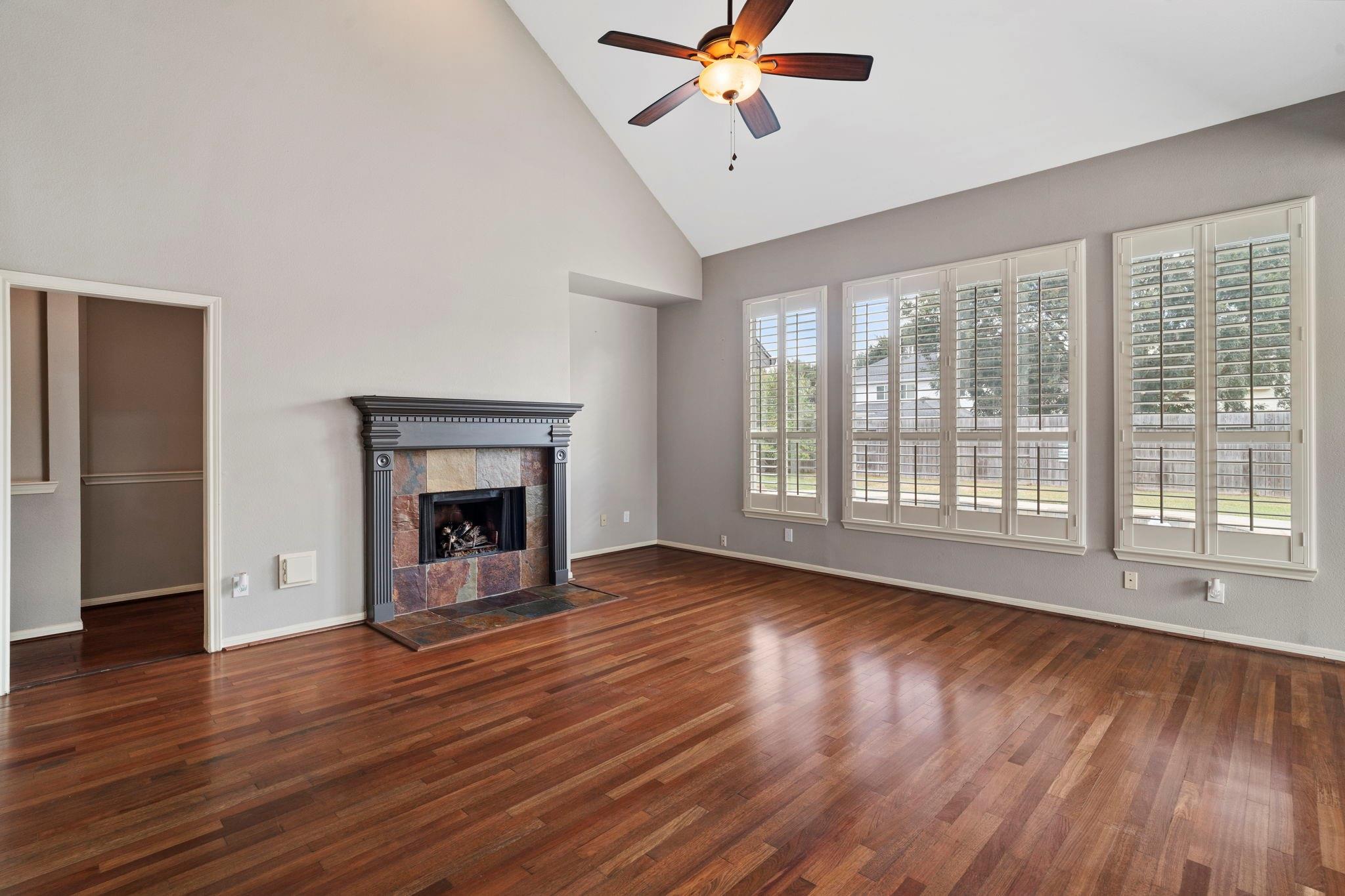 Empty living room with a stone fireplace, hardwood floors, and tall shuttered windows.