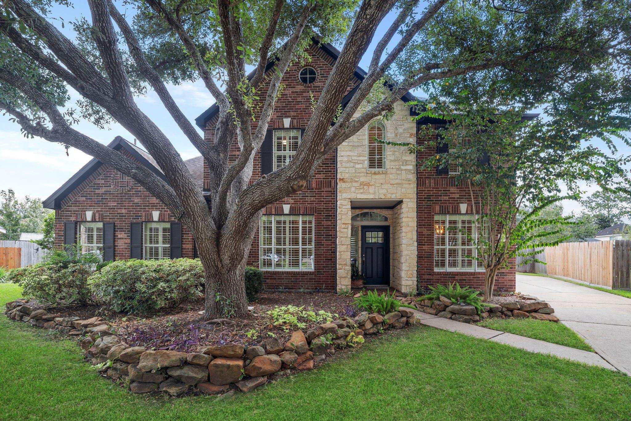 Two-story brick and stone home framed by a large shade tree, with a curved stone garden bed and side driveway.