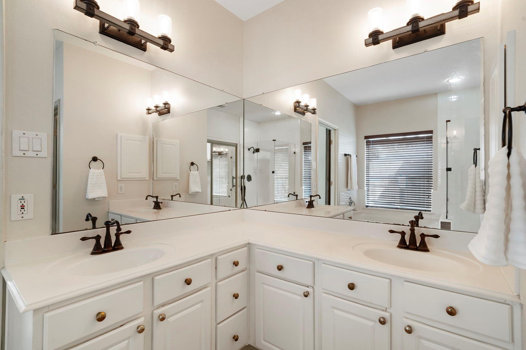 Bathroom double vanity with large mirrors reflecting a glass shower and window blinds.