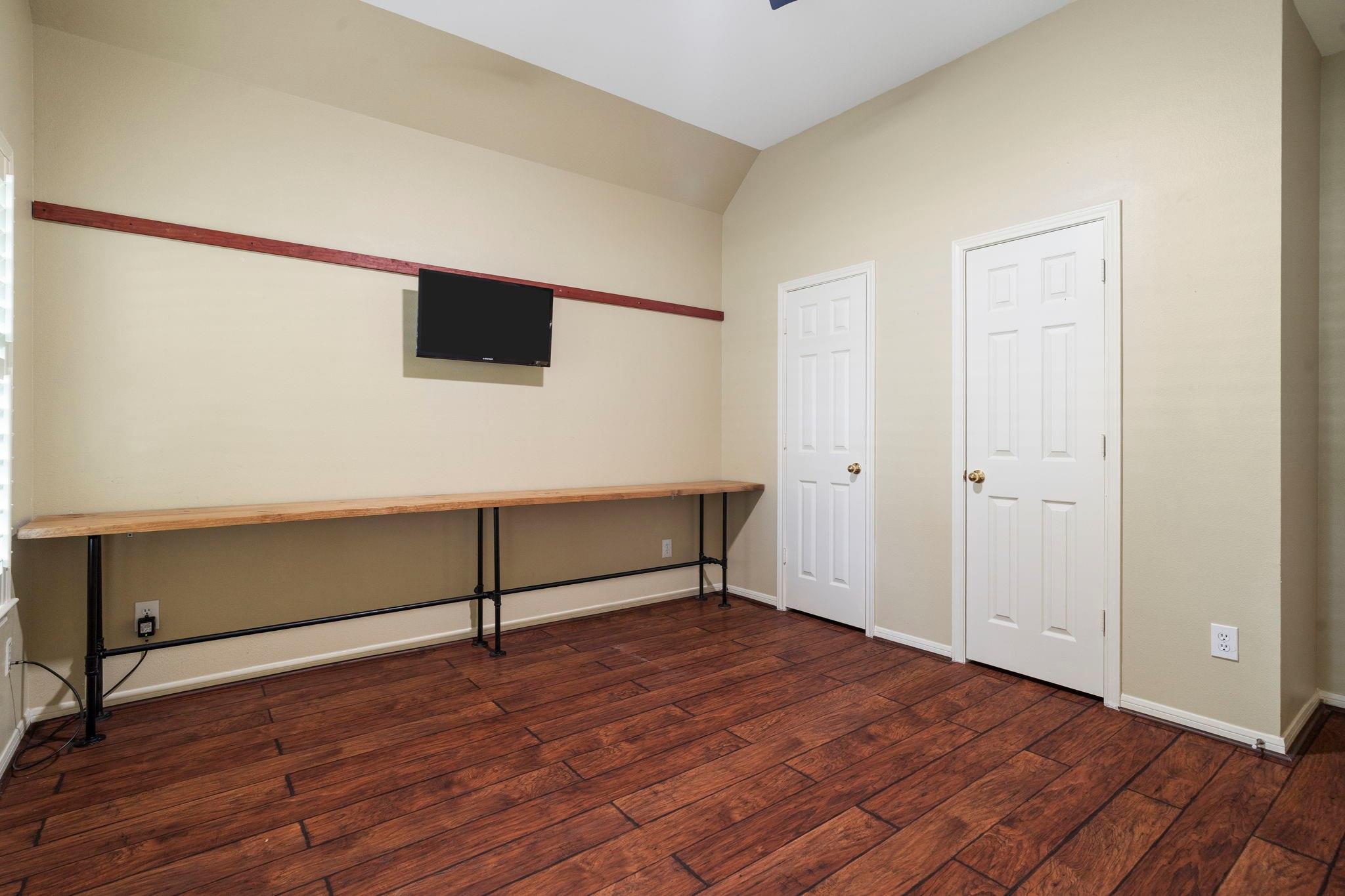 Interior room with a long workbench, wall-mounted TV, sloped ceiling, and dark wood-look flooring.