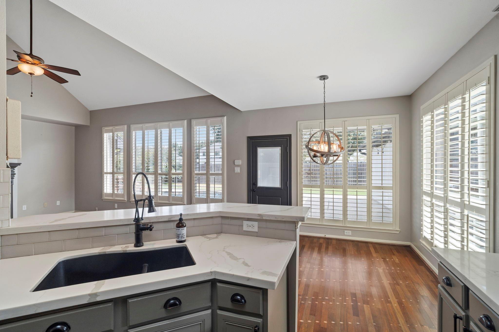 Bright kitchen with a stone island and dark sink, plantation shutters, and adjoining hardwood-floor area.