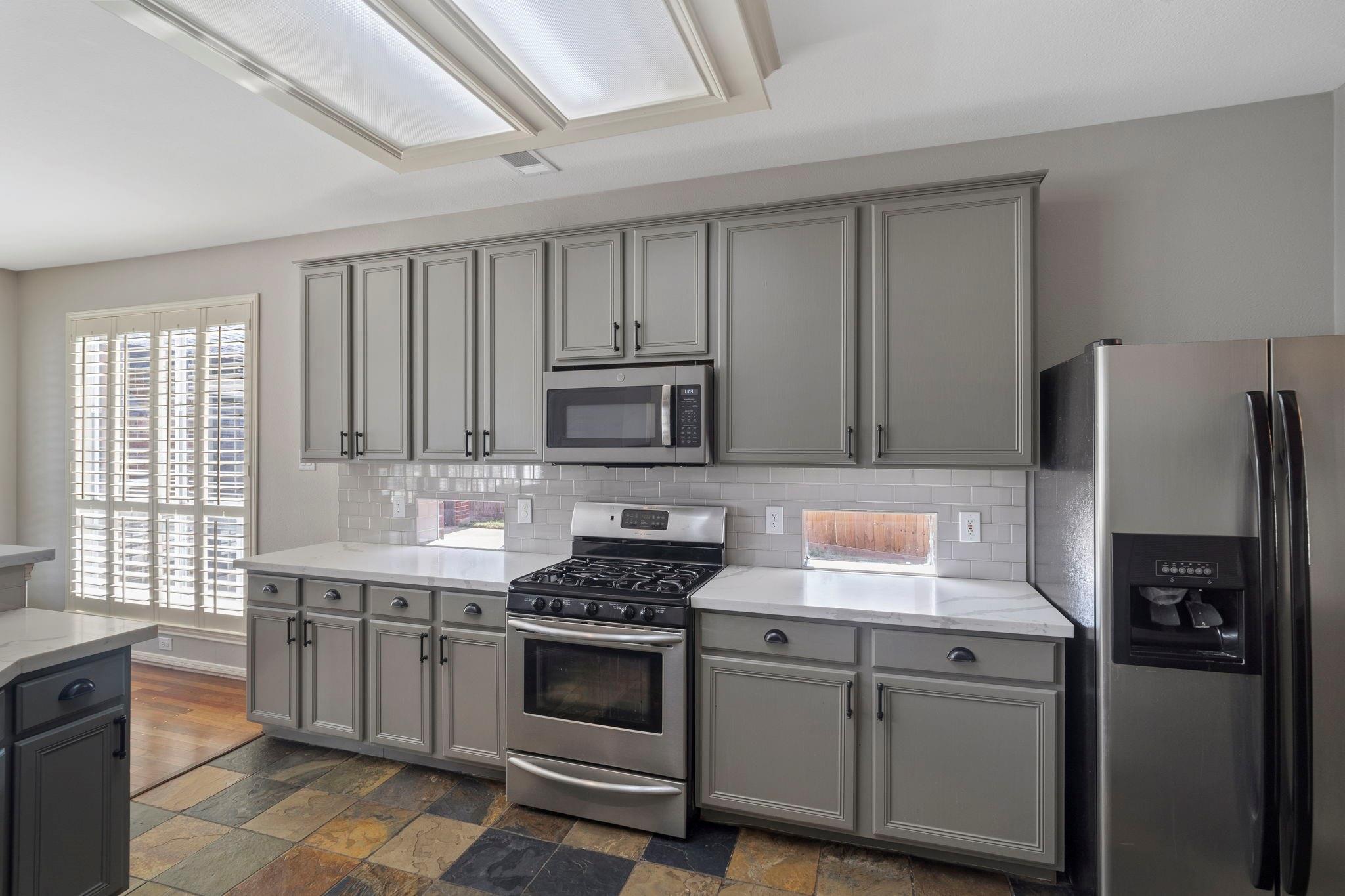 Kitchen with gray cabinets, stainless steel appliances, a large skylight, and multicolored stone tile flooring.