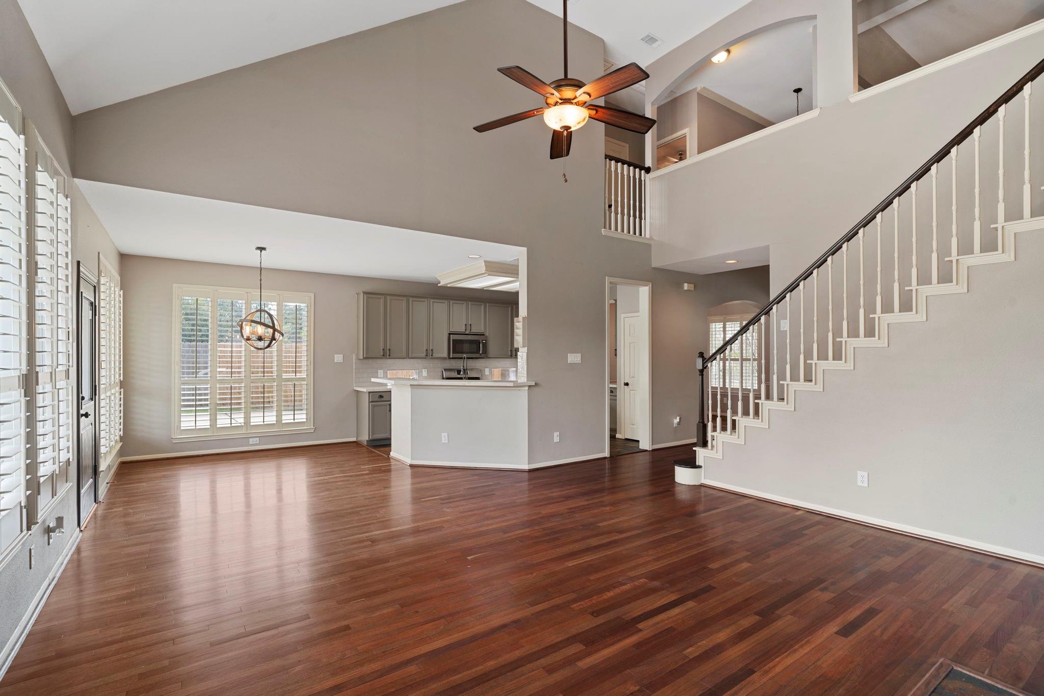 Open living area with glossy hardwood floors, high ceiling, plantation shutters, and staircase beside the kitchen.