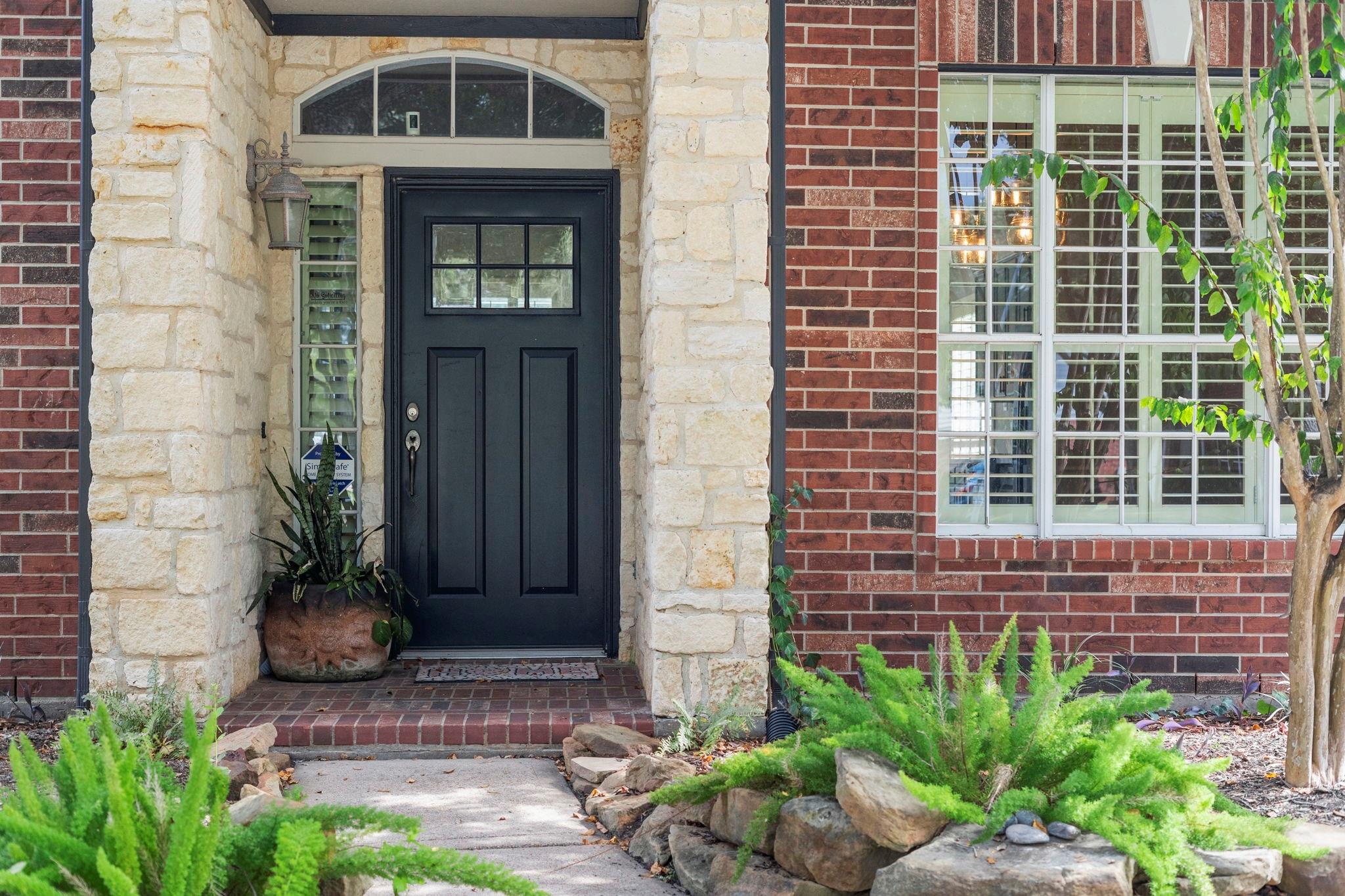 Brick home entrance with a black door framed by pale stone and a landscaped walkway.