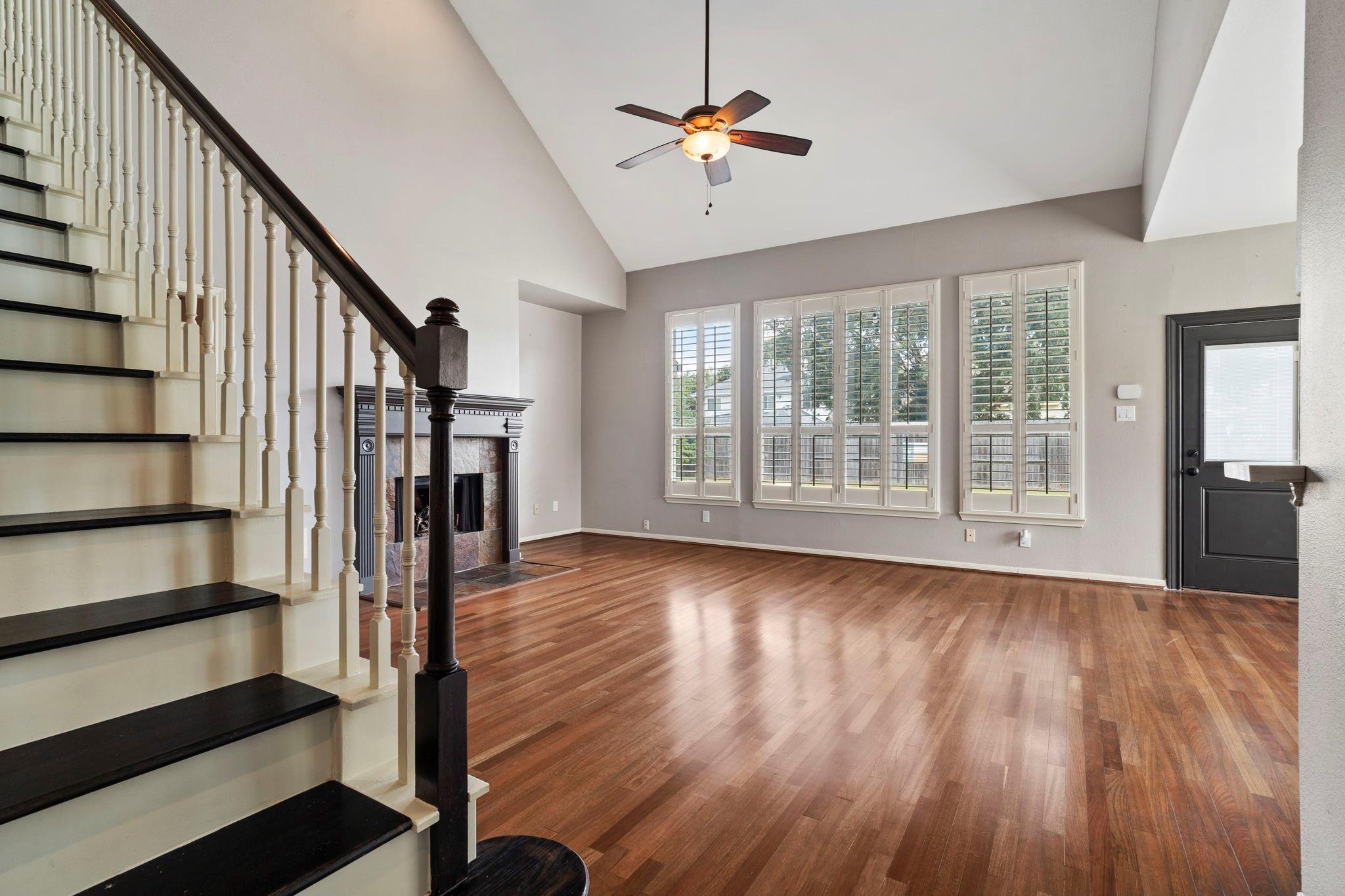 Empty living room with hardwood floors, fireplace, staircase, and shuttered windows.