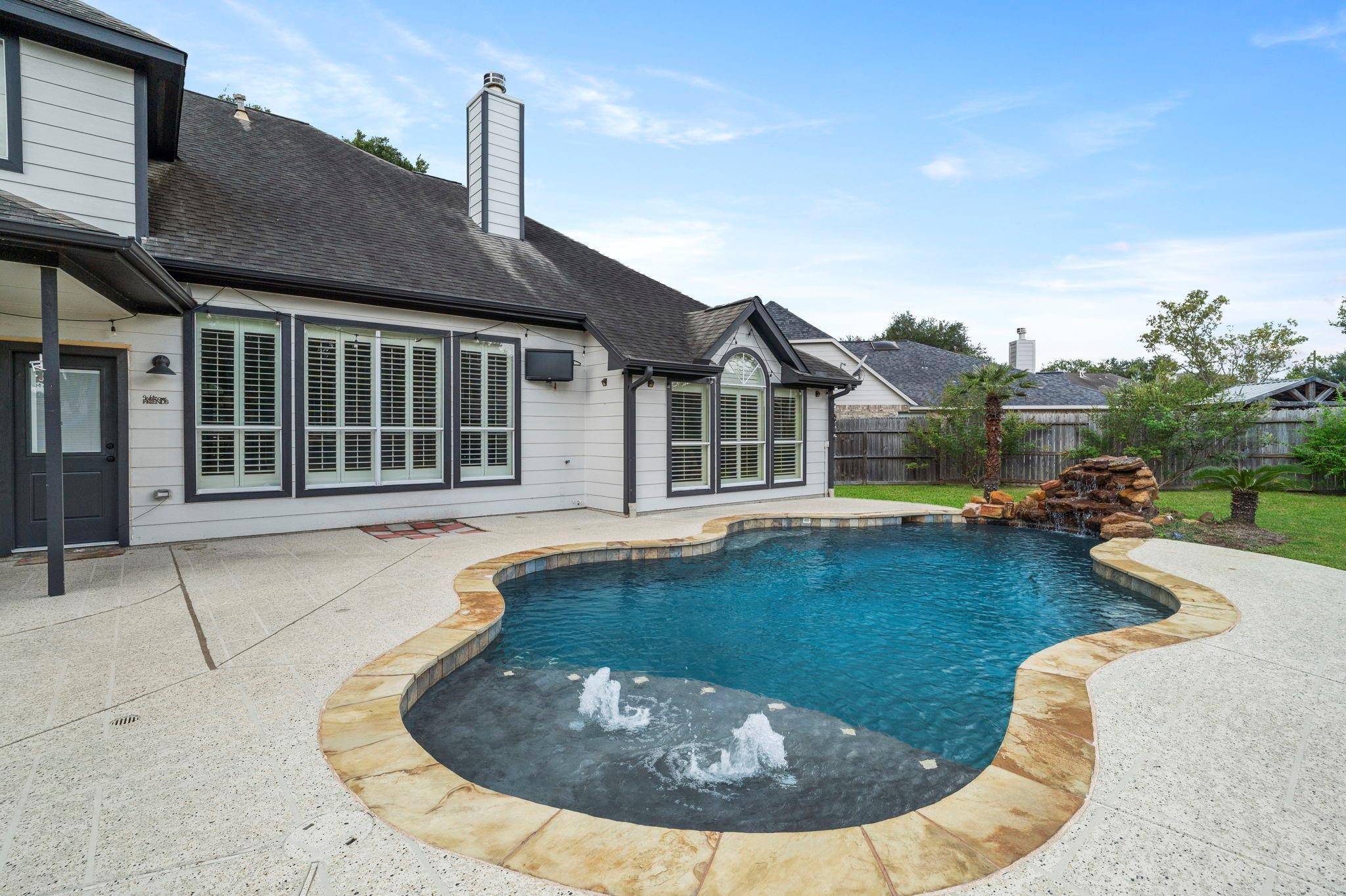 Rear of a light-colored house beside a curved pool with bubbling fountains and a rock waterfall.