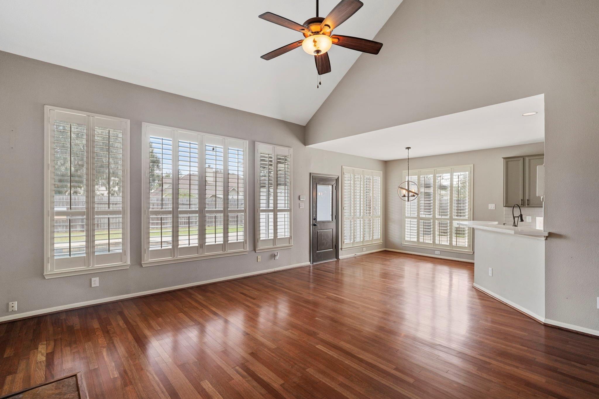 Vacant living and dining area with vaulted ceiling, dark wood floors, plantation-shuttered windows, and ceiling fan.