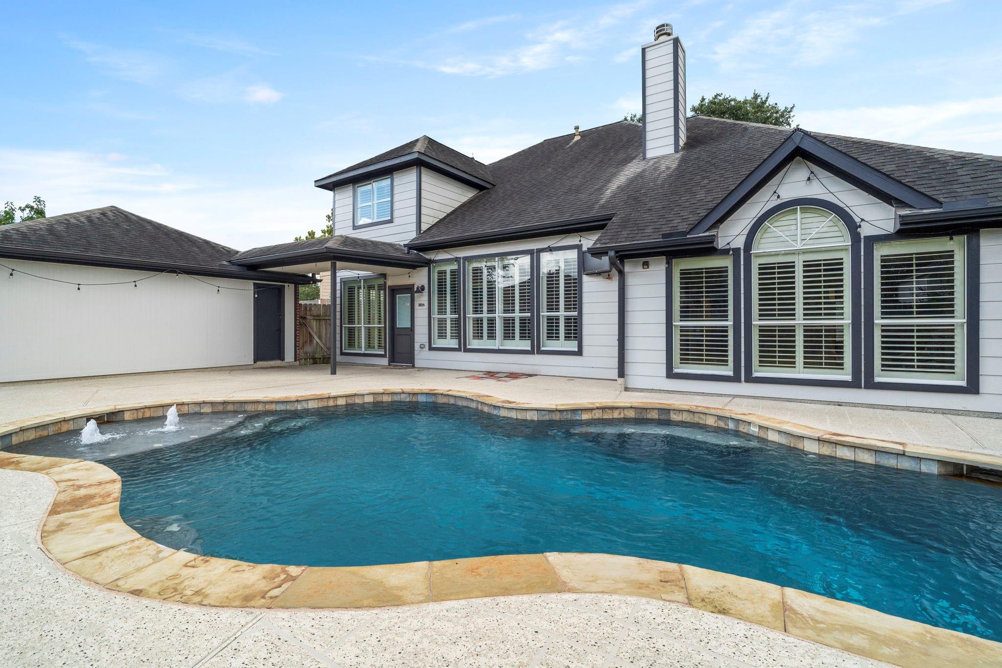 Rear of a gray two-story house beside a curved pool with fountains and a spacious patio.