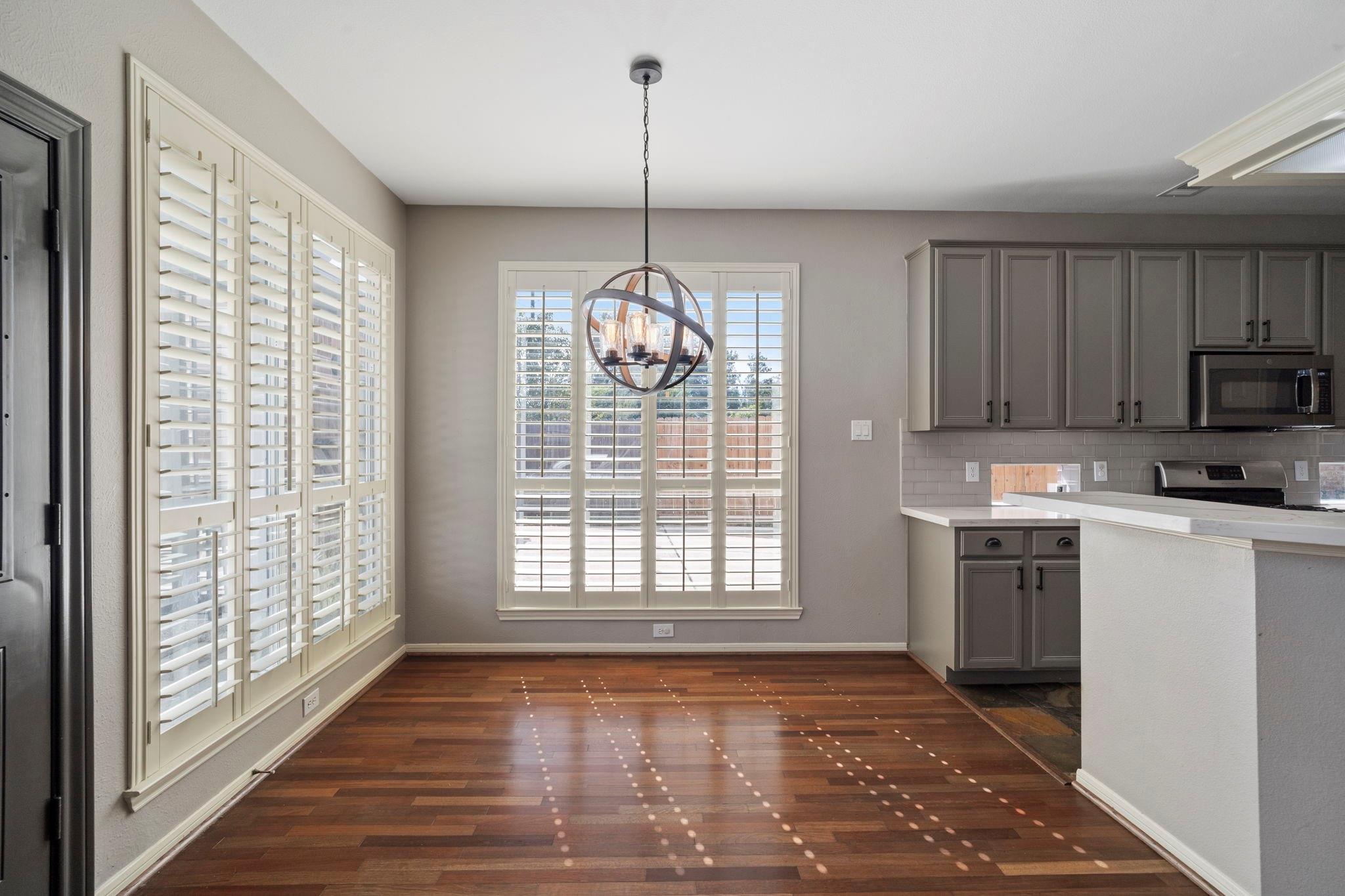 Vacant dining area with hardwood floors, plantation shutters, and an orb chandelier beside the kitchen.
