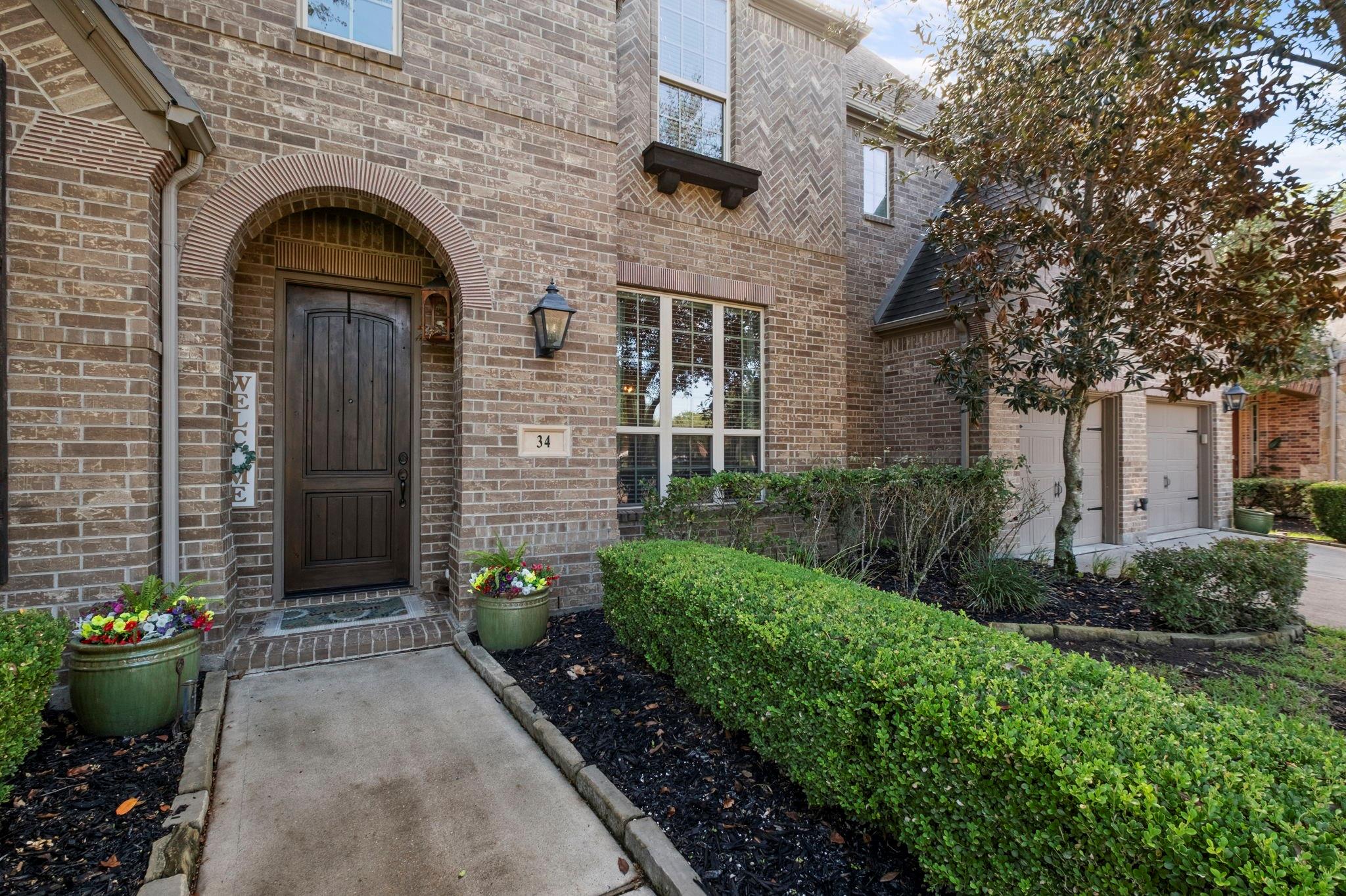 Two-story brick home with an arched entry, dark wood front door, flower planters, and trimmed hedges.