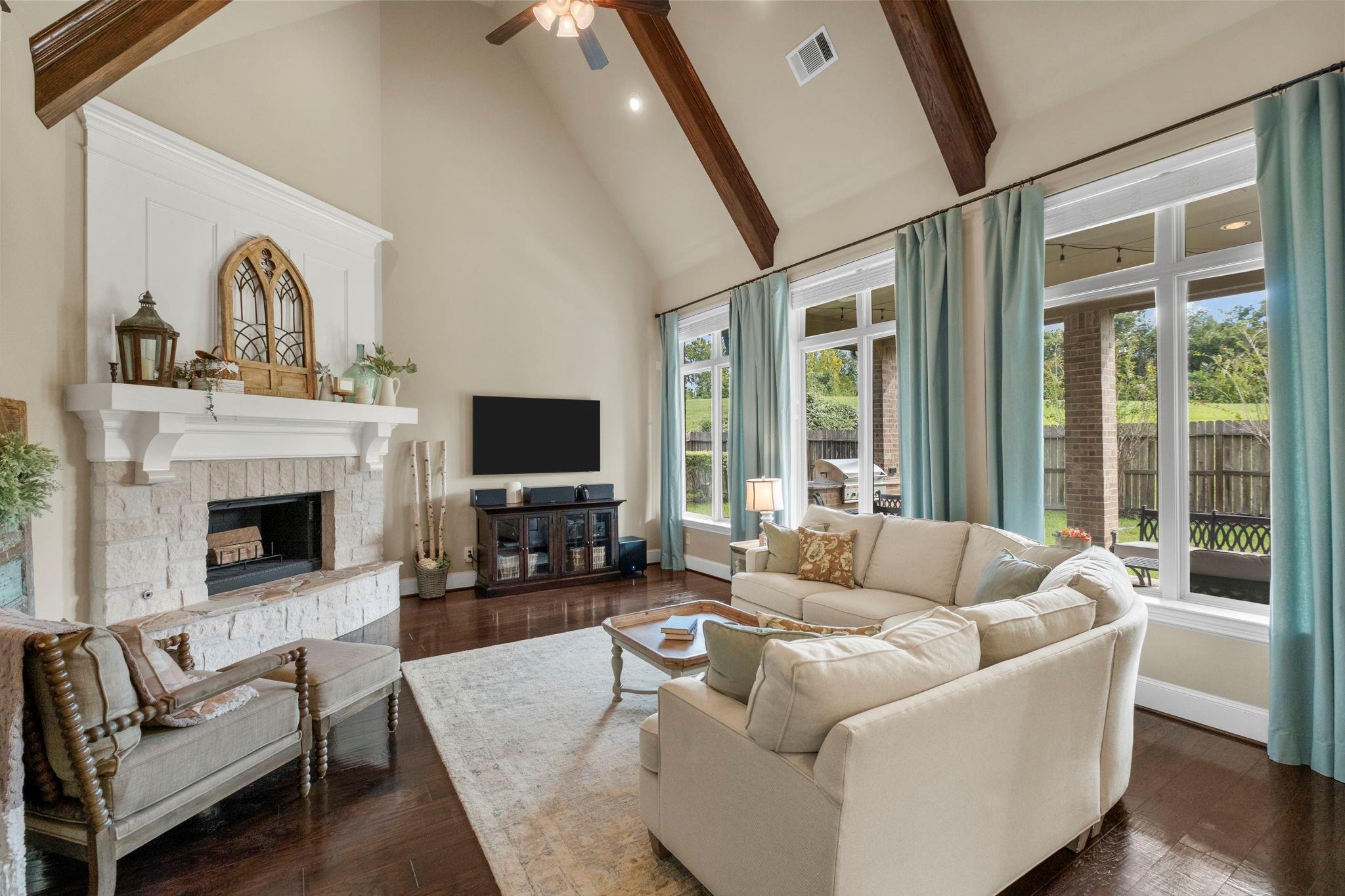 Living room with a stone fireplace, vaulted wood-beam ceiling, and cream sectional beside tall windows.