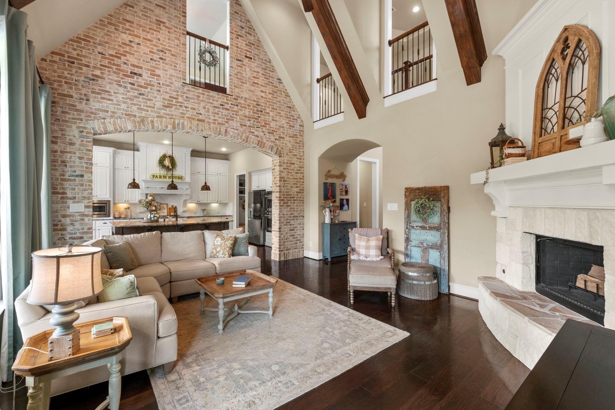 Open living room with vaulted wood-beam ceiling, brick kitchen arch, beige sectional, and stone fireplace.