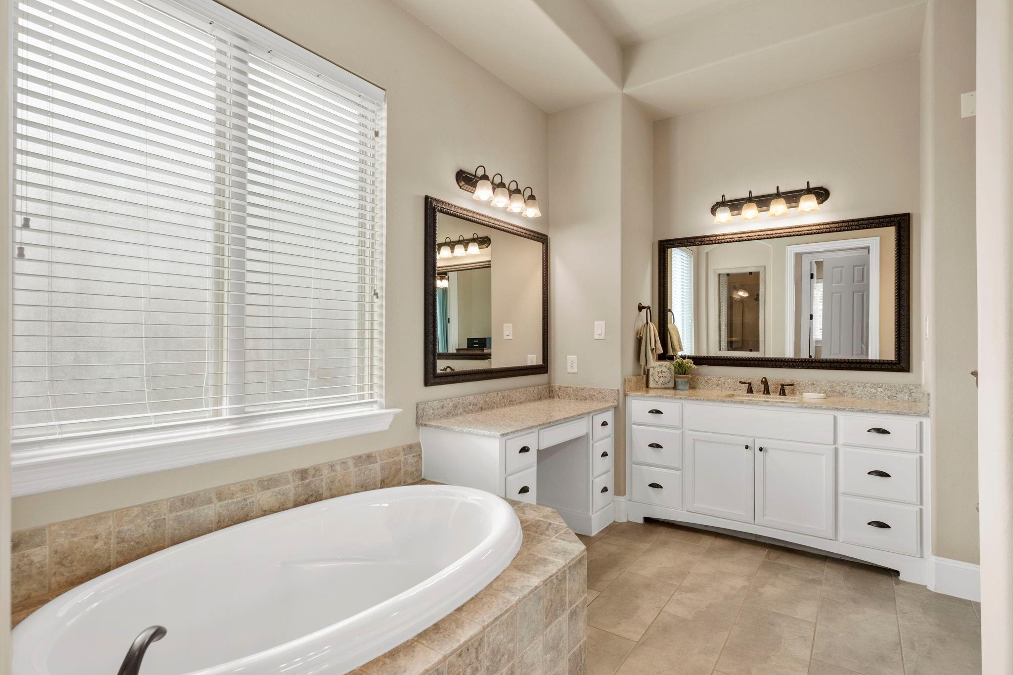 Primary bathroom with a tiled soaking tub, white vanities, stone counters, and large mirrors.
