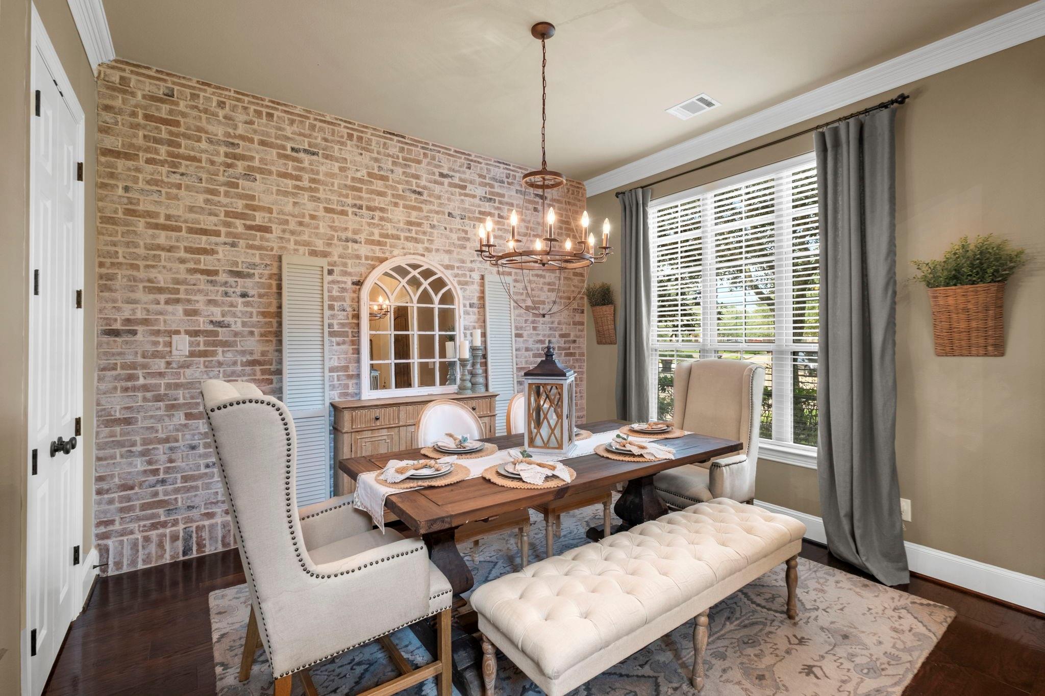 Dining room with a brick accent wall, long wooden table, upholstered seating, and chandelier.