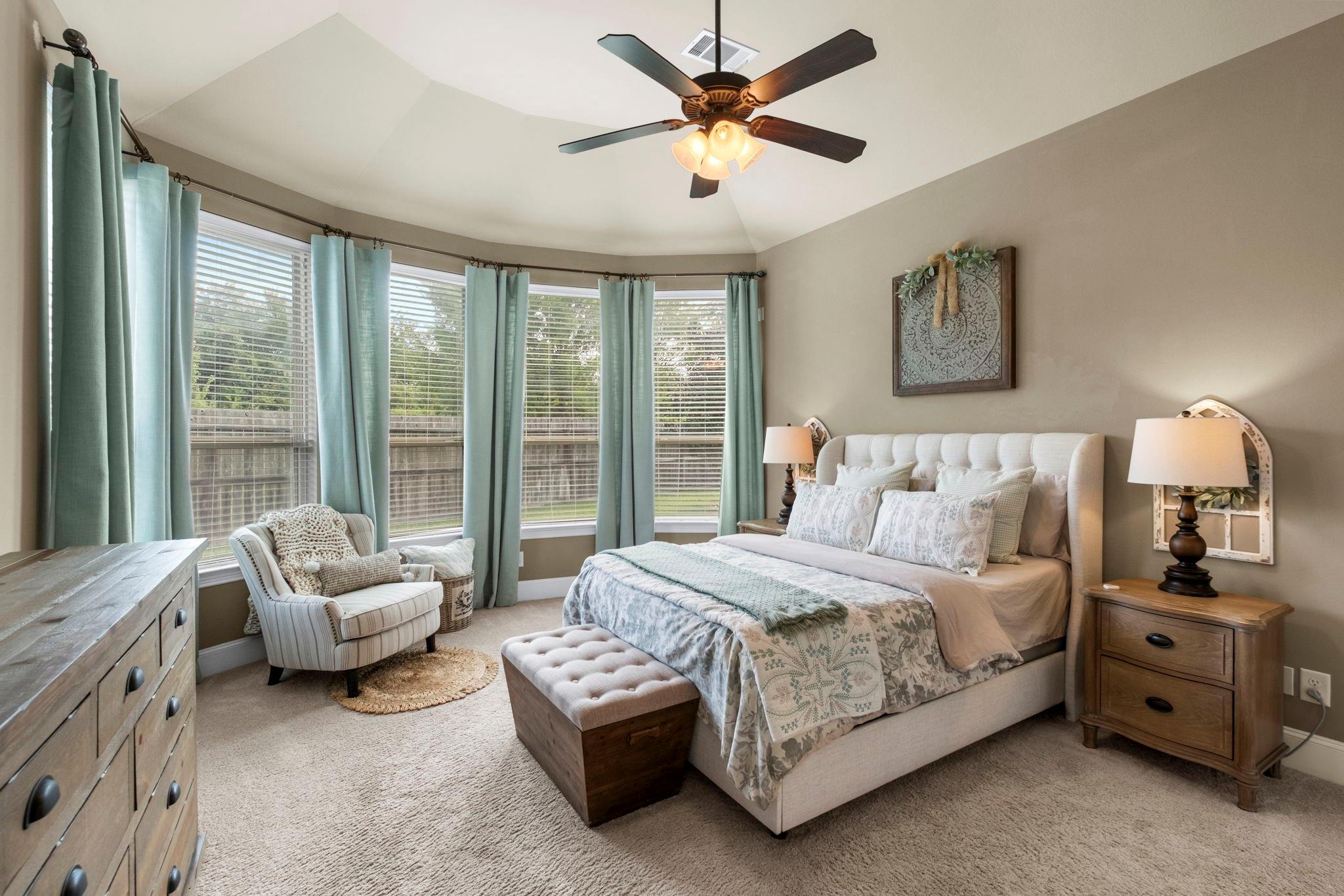 Furnished bedroom with a large bed, blue-green curtains, wide windows, and an upholstered armchair.