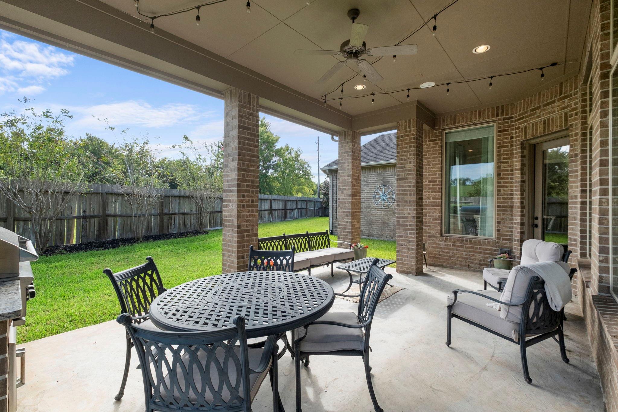 Covered brick patio with outdoor dining furniture and views of a fenced backyard.