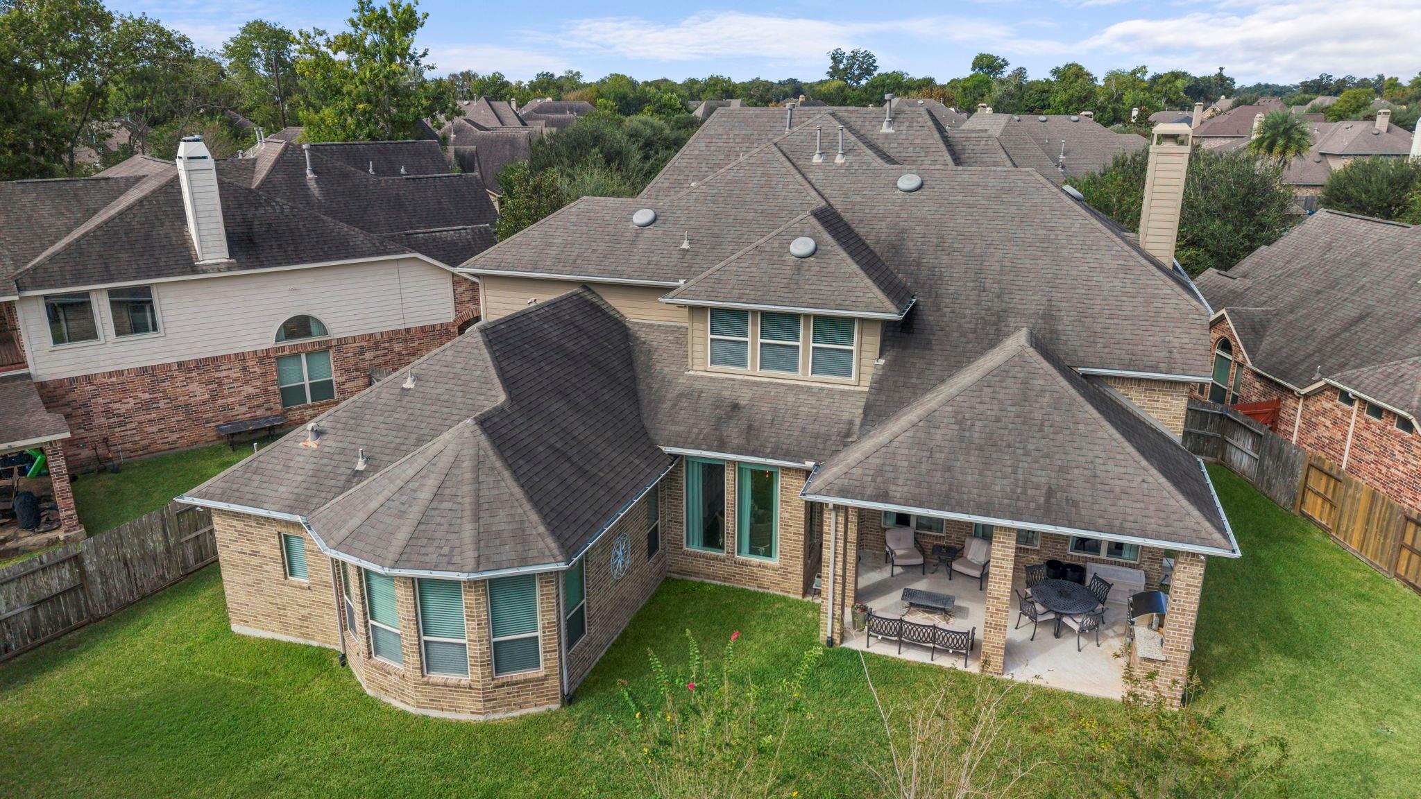 Aerial view of a brick home’s rear roof, covered patio, and fenced grassy backyard.