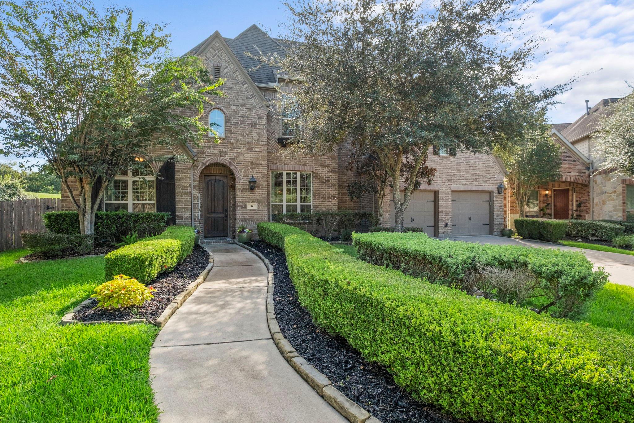 Two-story brick house with an arched entry, double garage, and curved walkway through trimmed hedges.