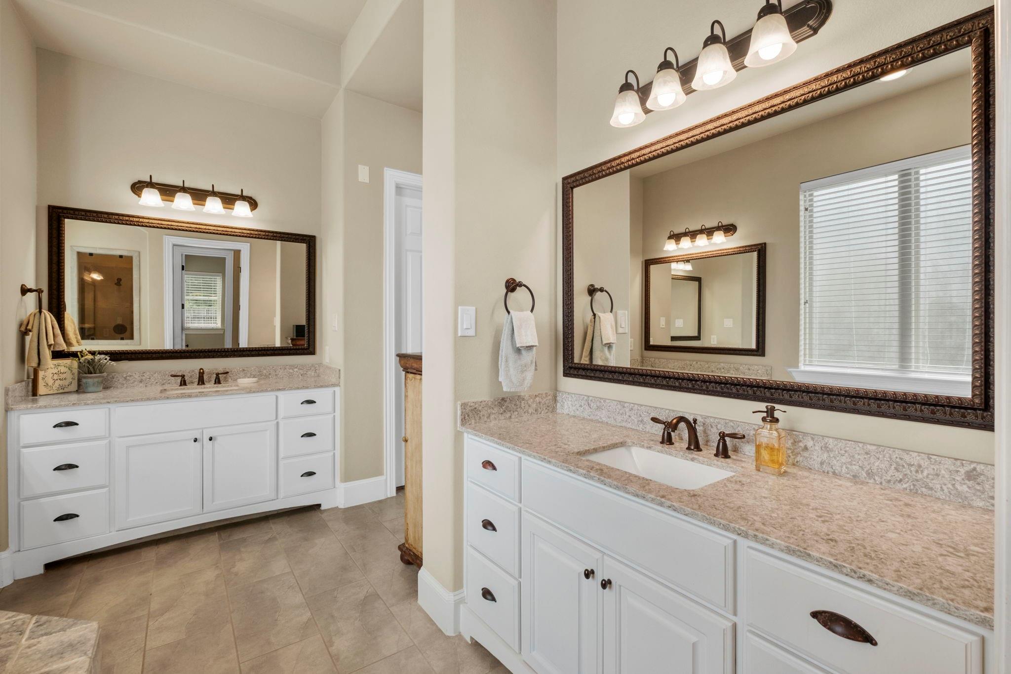 Bathroom with two white vanities, stone countertops, large framed mirrors, and a window with blinds.