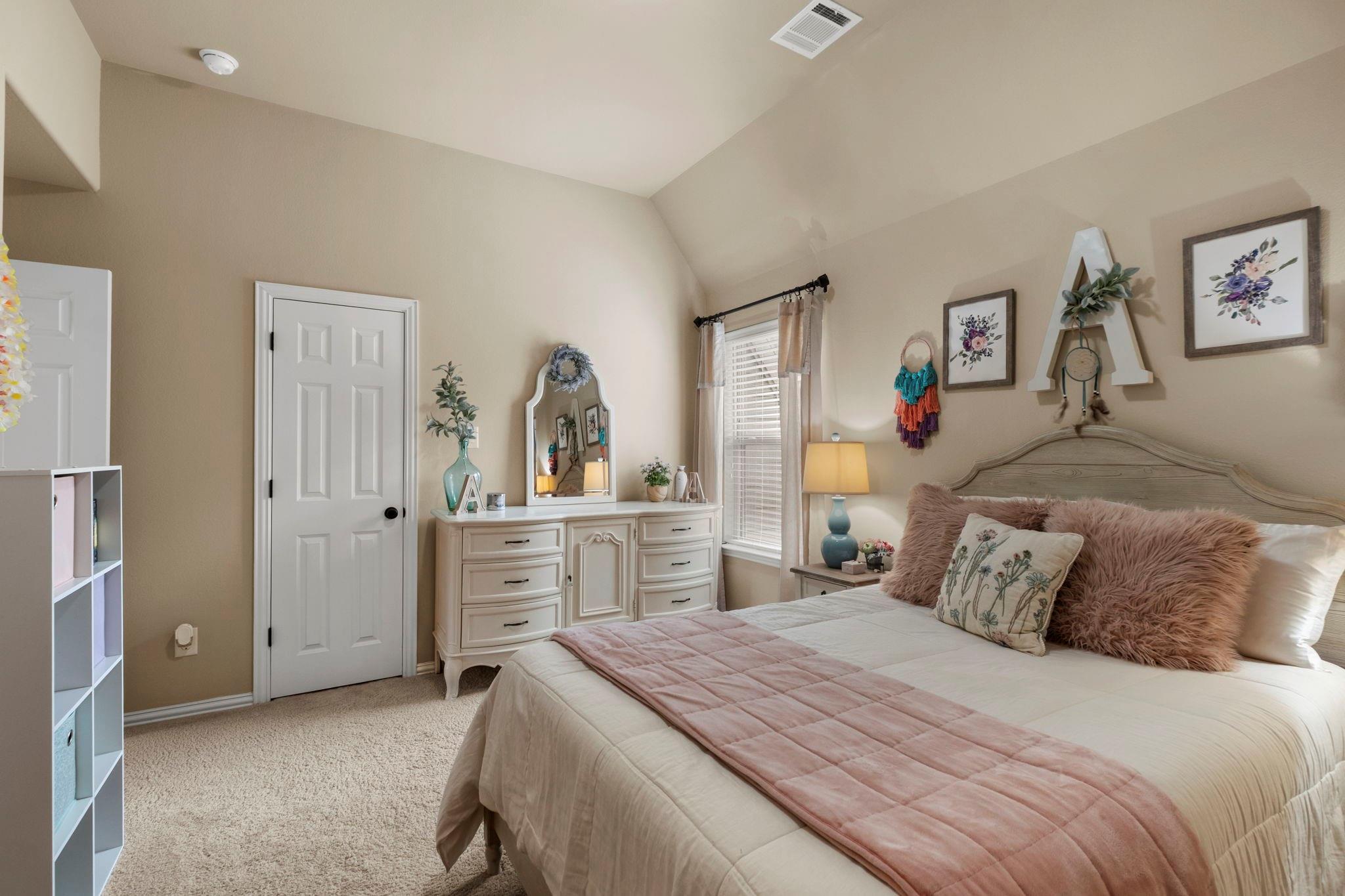 Furnished bedroom with a vaulted ceiling, pink quilt and pillows, and a white dresser beside the window.